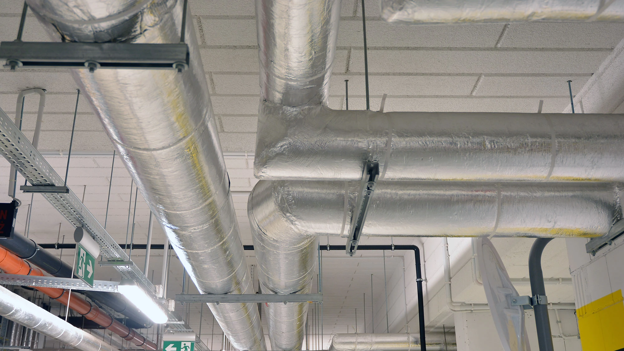 Ceiling view of silver insulated pipes and ventilation ducts in an industrial or commercial building.