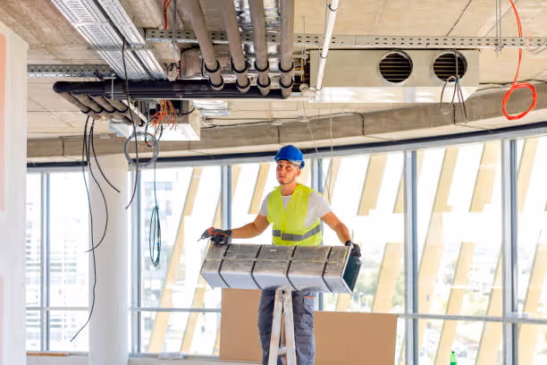 Construction worker wearing a blue helmet and yellow safety vest installing ductwork in a building under construction.