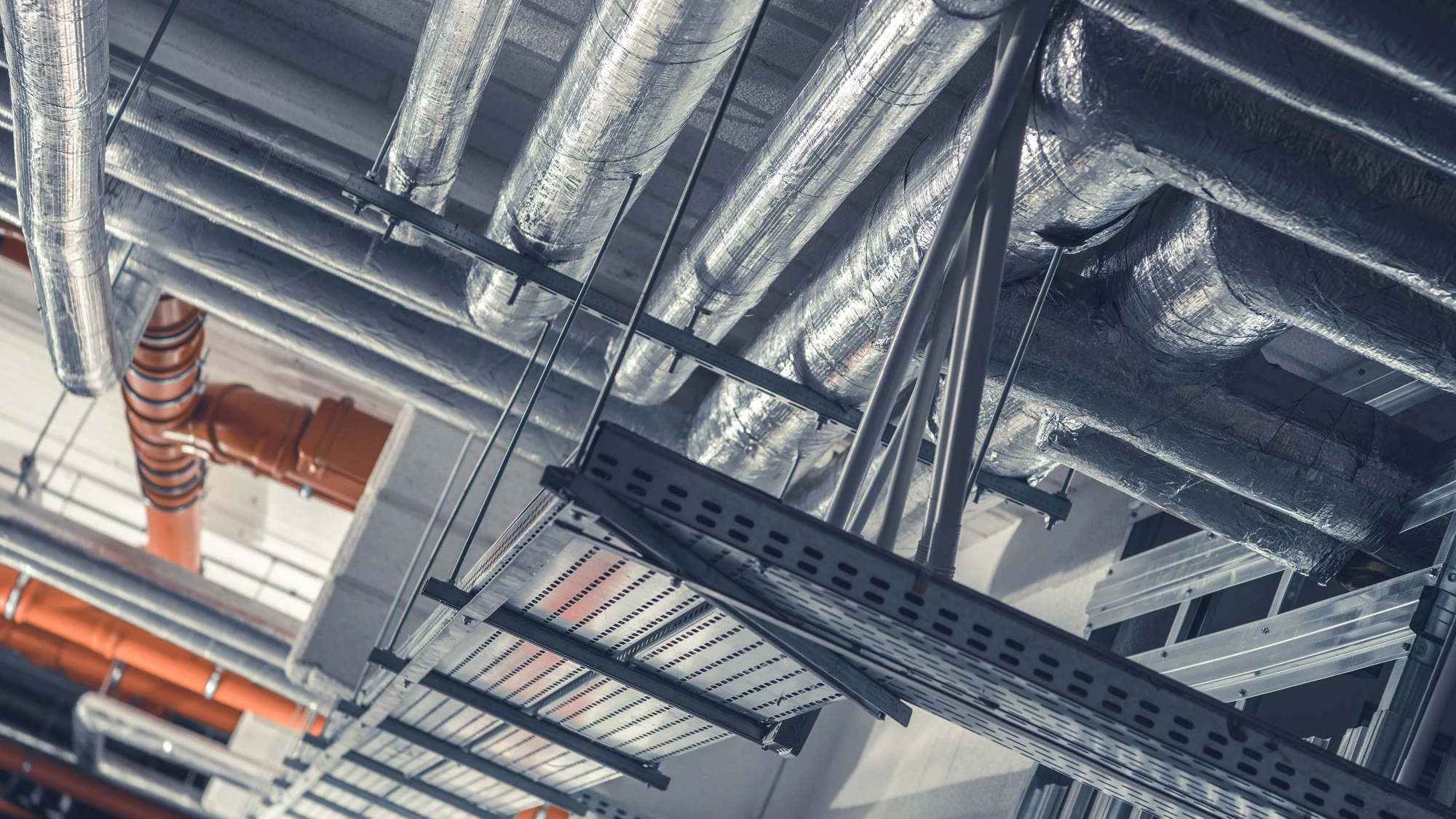 Ceiling with insulated silver HVAC ducts and orange pipes in an industrial building.