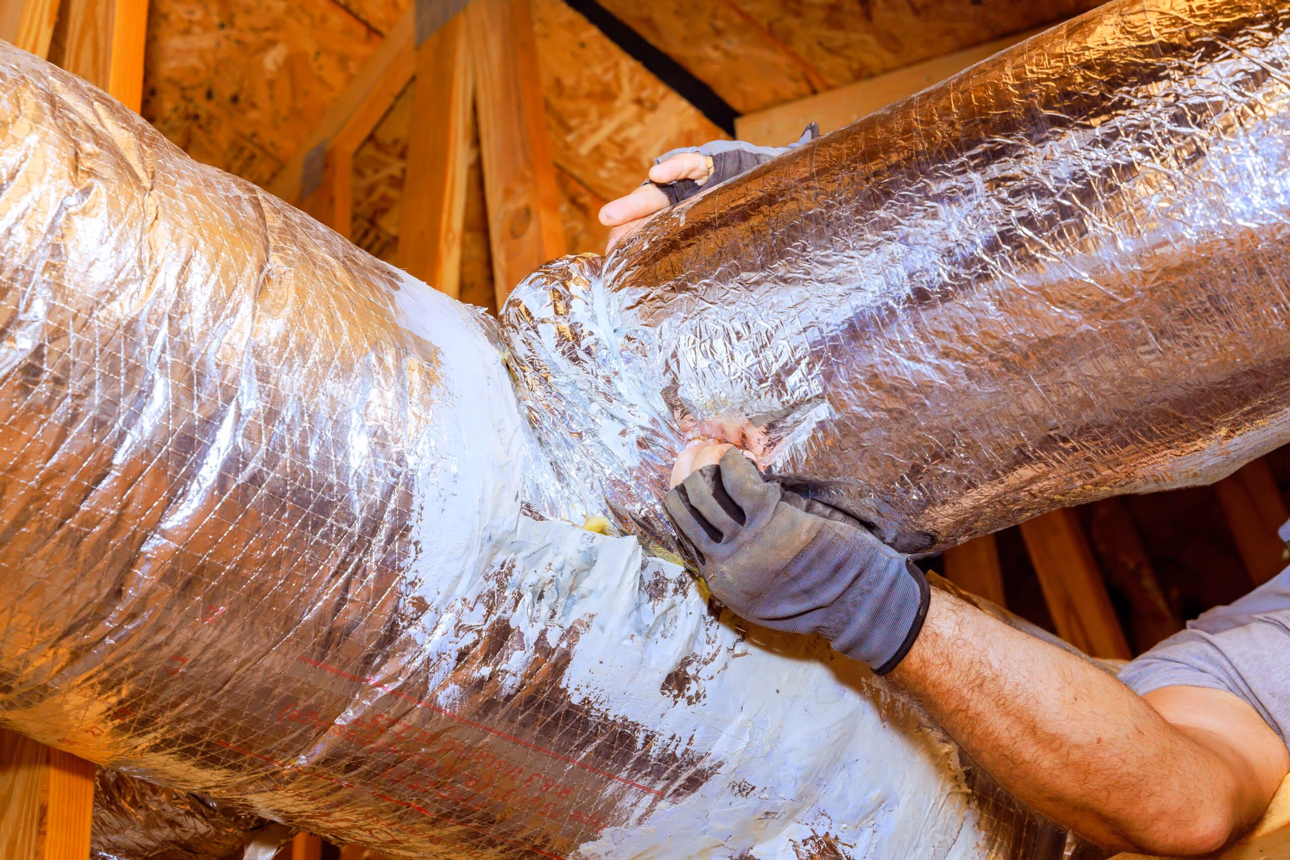 Person wearing gloves sealing a shiny insulated air duct in an attic with wooden beams.