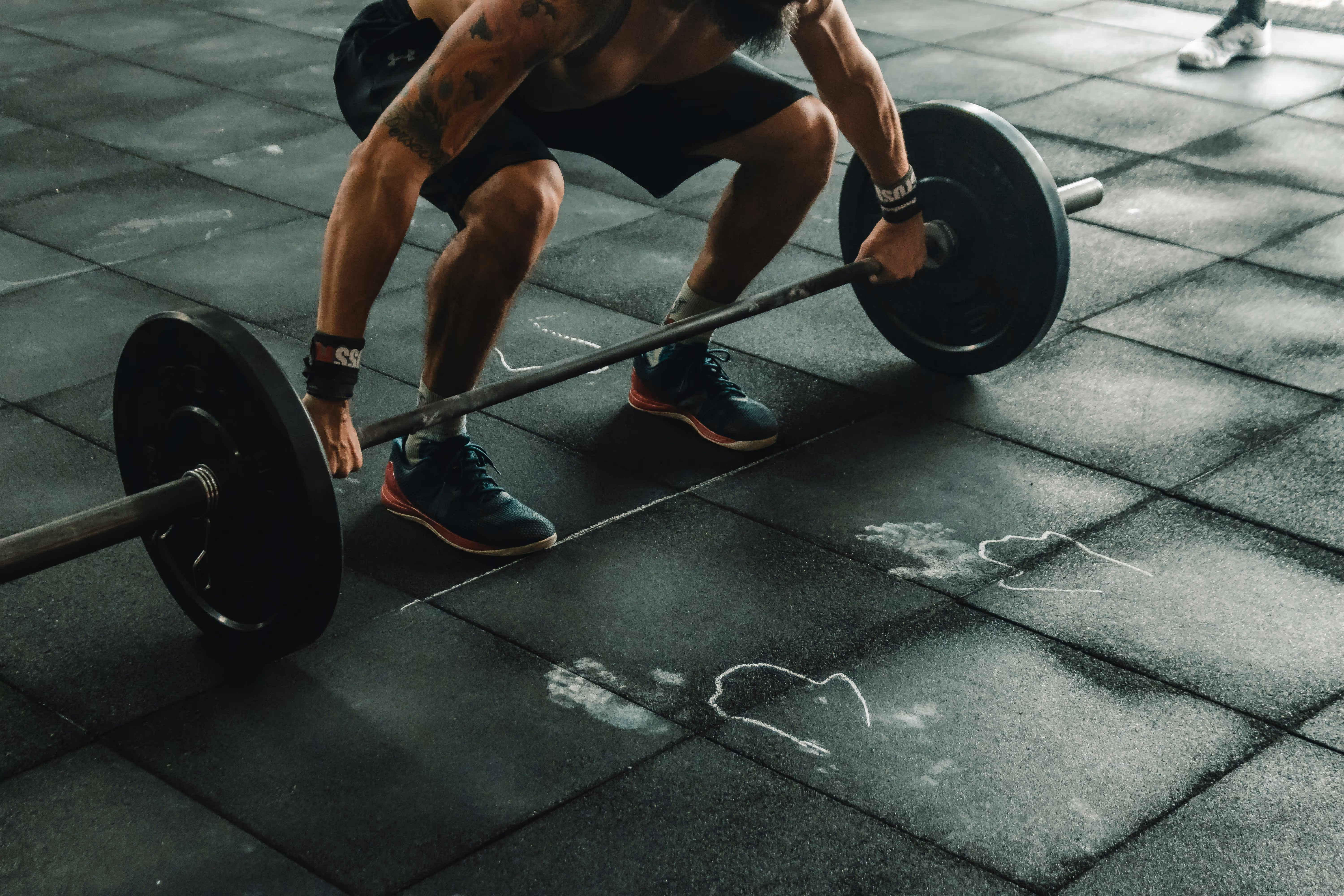 Man with tattoos and wrist wraps preparing to lift a barbell on a gym floor with chalk footprints.