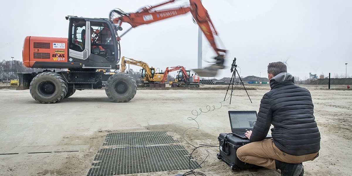 Man in a black jacket kneeling on dirt next to a laptop conducting acoustic noise monitoring of an orange excavator in an open construction site.
