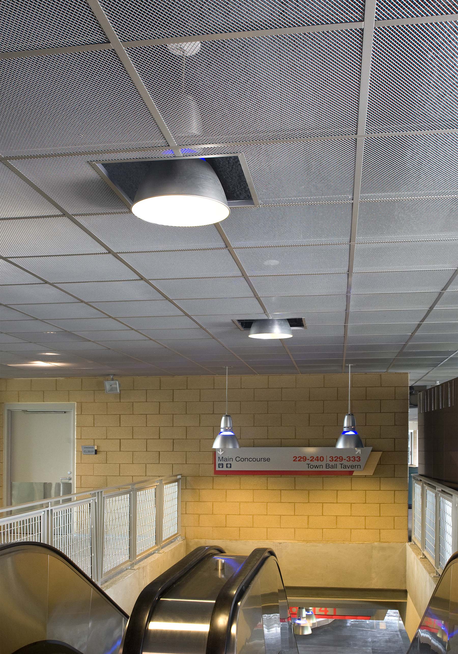Indoor escalators descending with beige brick walls, metal mesh ceiling, and a sign indicating Main Concourse and Town Ball Tavern directions.