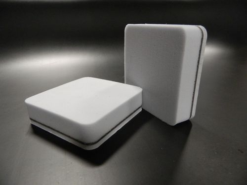 Two square, white foam pads with rounded edges and a black layer in the middle, placed on a dark surface.