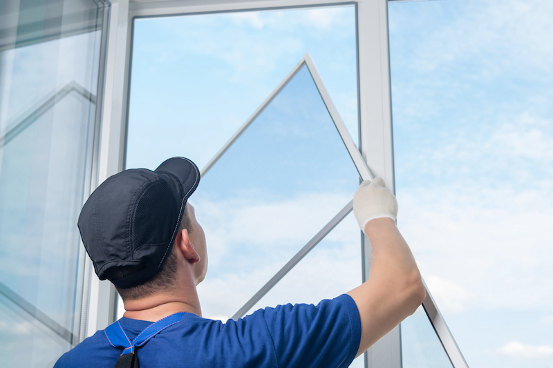 Worker in blue shirt and cap installing or removing a triangular window panel with glass against a blue sky background.