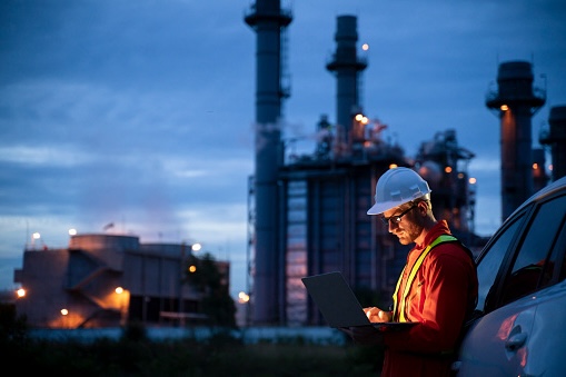 Engineer in a hard hat and safety vest using laptop near a car with an industrial plant lit up at dusk in the background.