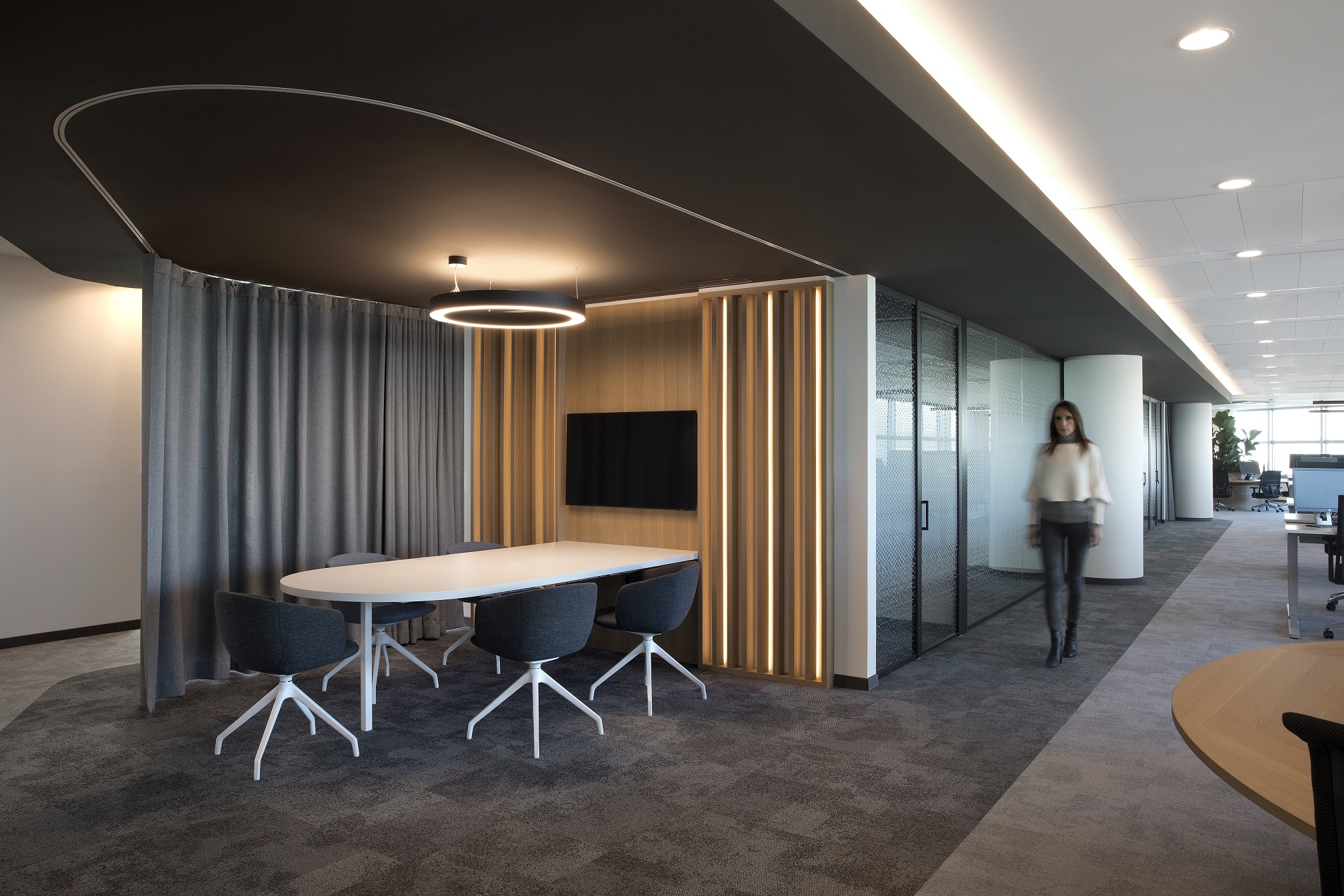 Modern office meeting area with a white table, five dark grey chairs, wood panel with vertical light strips, and a circular ceiling light, adjacent to a hallway where a person walks.