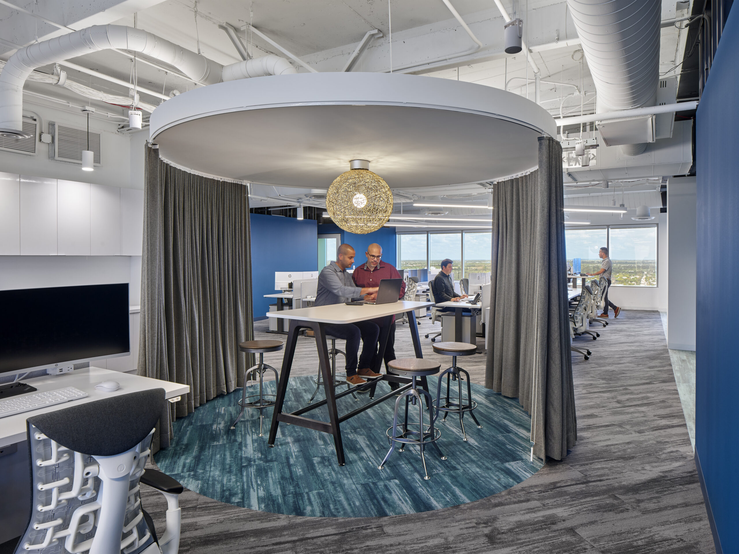 Modern office space with two men collaborating at a high table under a round ceiling fixture and four stools, surrounded by gray curtains and blue carpet, with other workers at desks and large windows in the background.