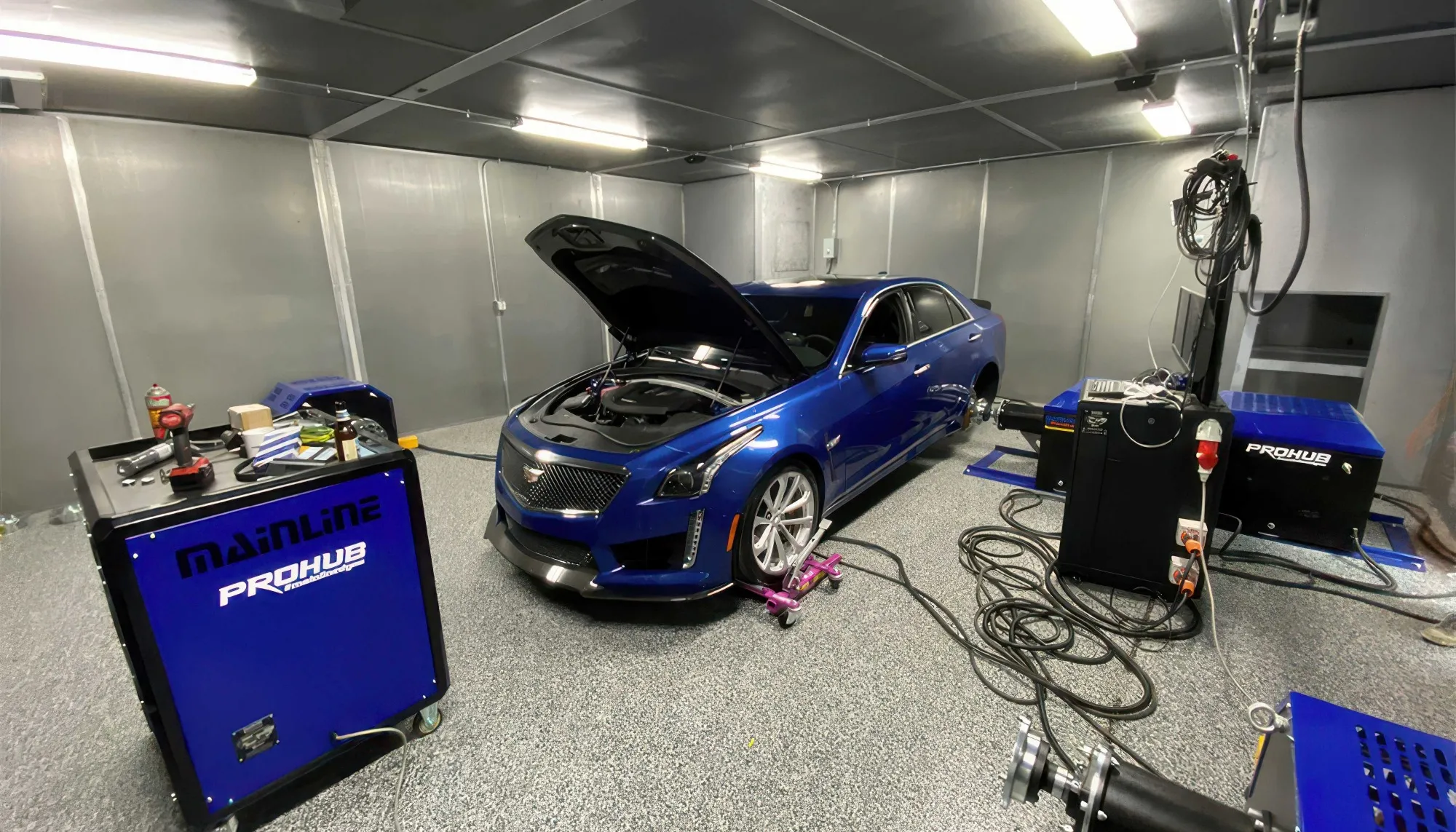Blue Cadillac sedan with open hood on a chassis dynamometer testing setup in a gray-walled, well-lit test cell.