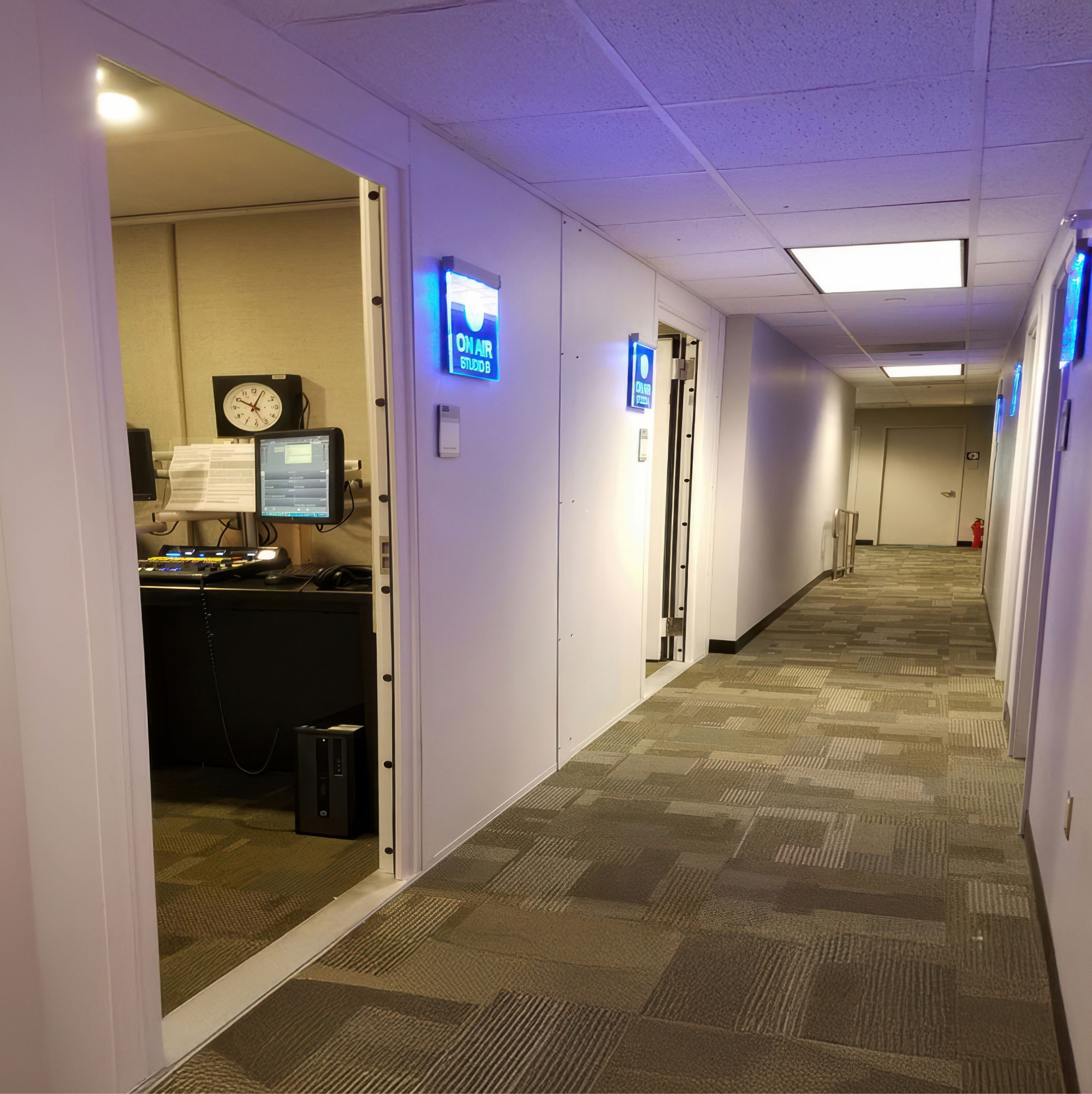 Hallway with carpeted floor and doors to rooms, one door open revealing a studio control room with computer monitors and an analog clock showing 9:10.