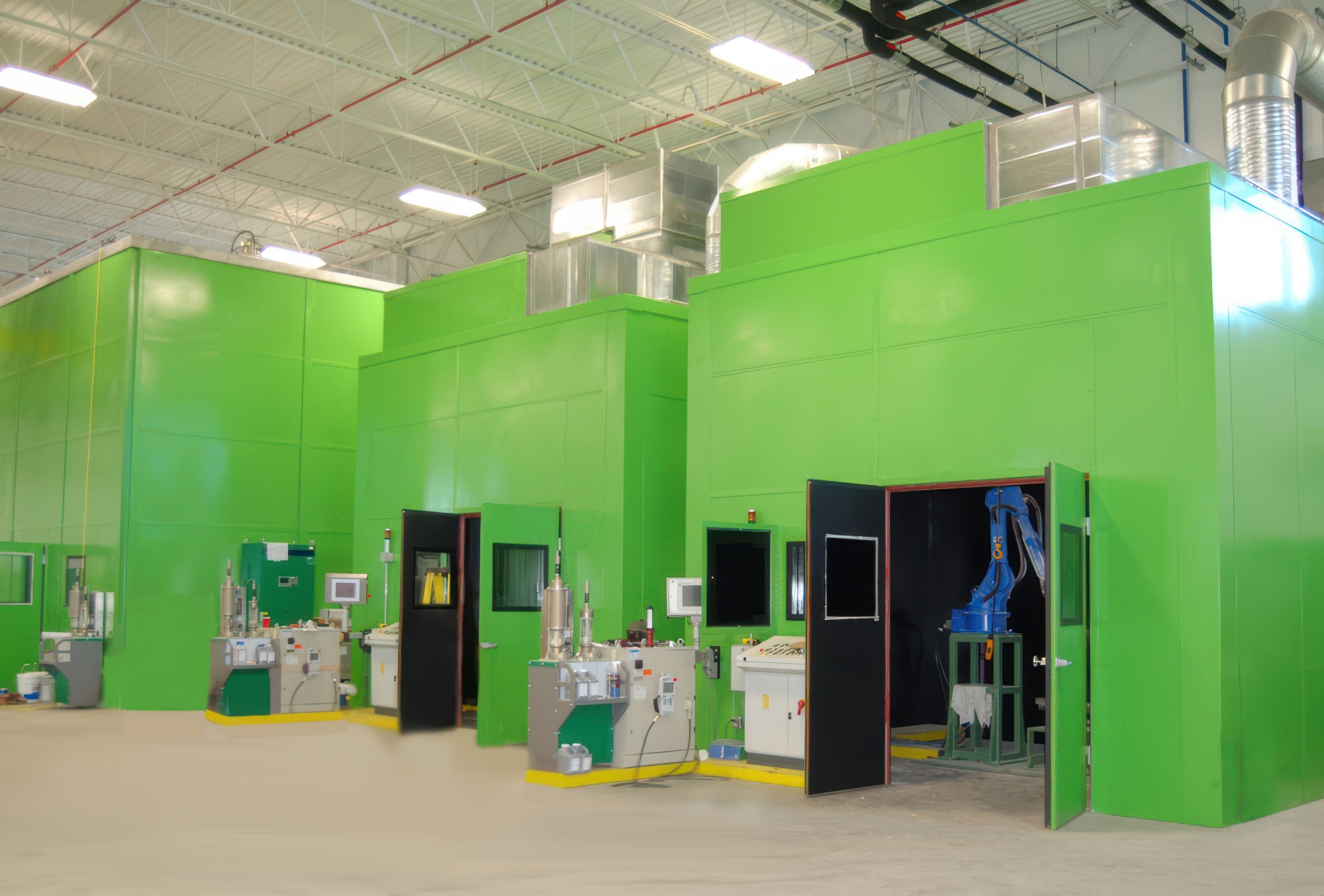 Industrial workspace with multiple large green modular rooms, each equipped with control panels and machinery, one room has a blue robotic arm inside.