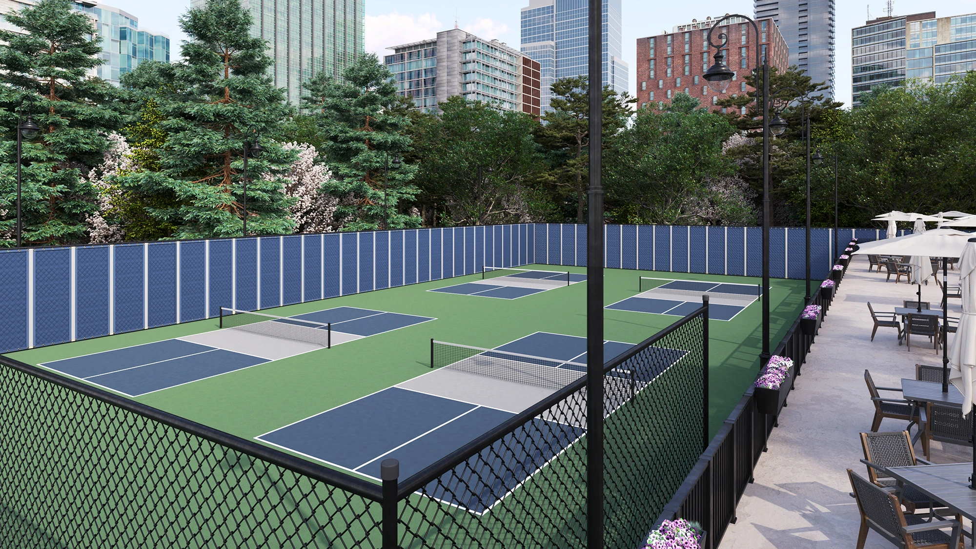 Four pickleball courts with green and blue surfaces surrounded by a black chain-link fence and an outdoor seating area with tables and umbrellas, set against a backdrop of urban buildings and trees.