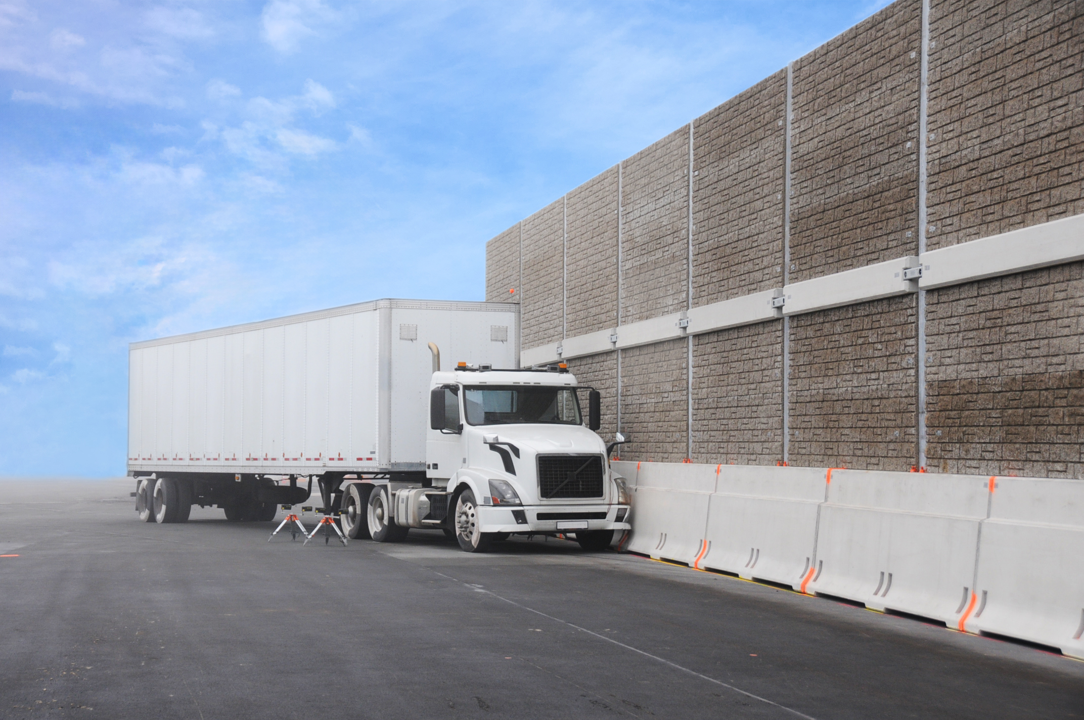 White semi-truck with trailer parked beside a concrete barrier wall under a blue sky.