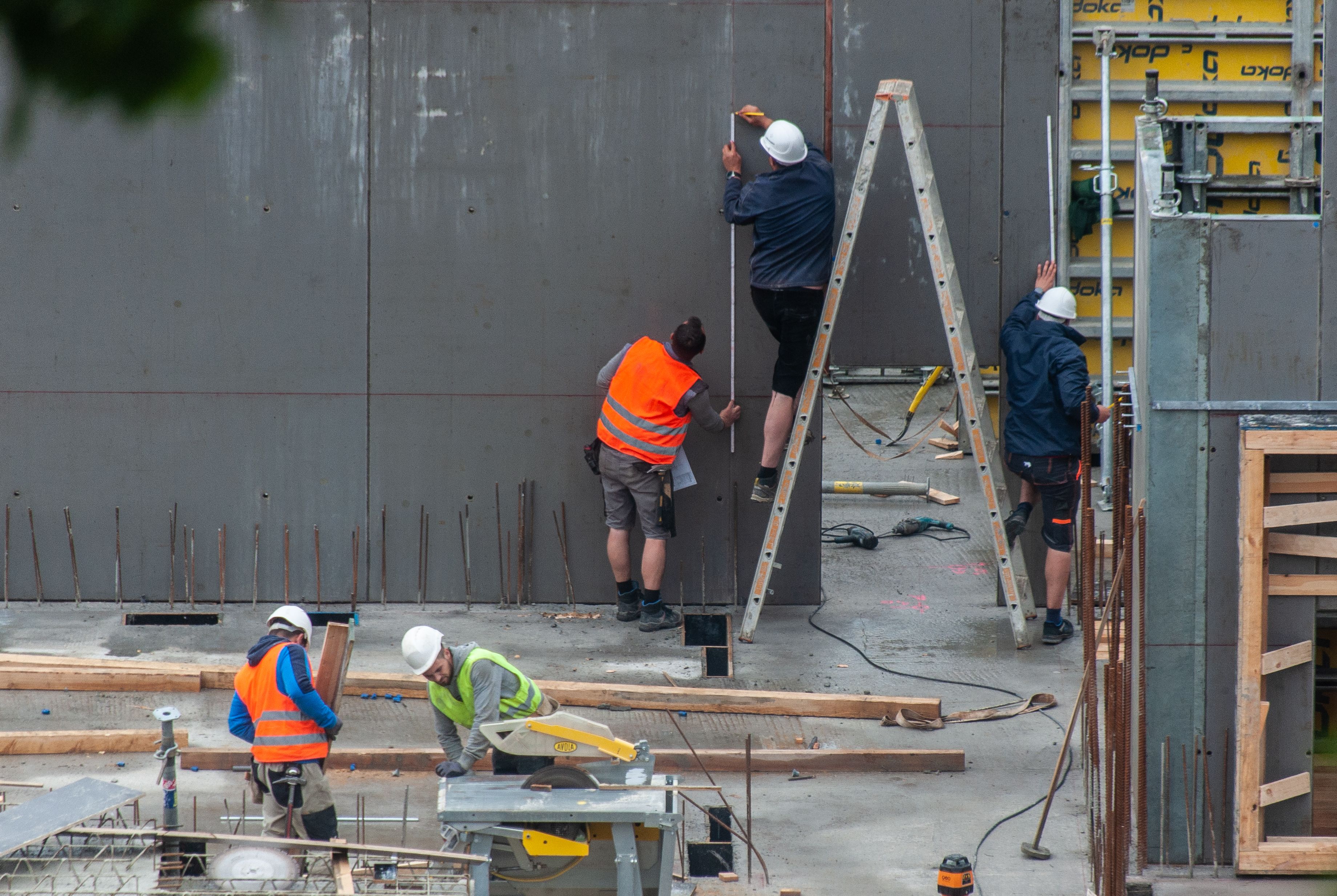 Construction workers in helmets and vests measuring and working on concrete walls and floor at a site.