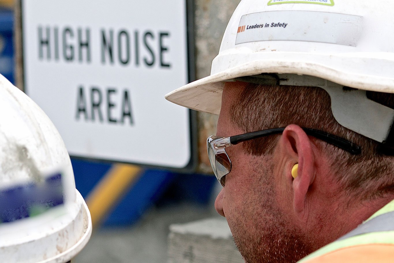Plant worker in a hard hat wearing a foam earplug and safety glasses beside a High Noise Area warning sign.