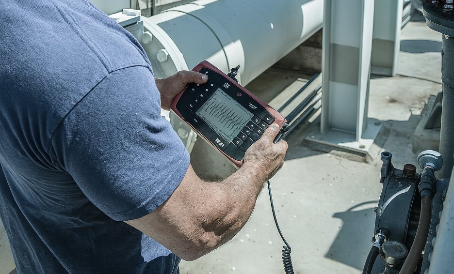 Technician taking an in-field sound reading with a handheld analyzer beside industrial piping and equipment.