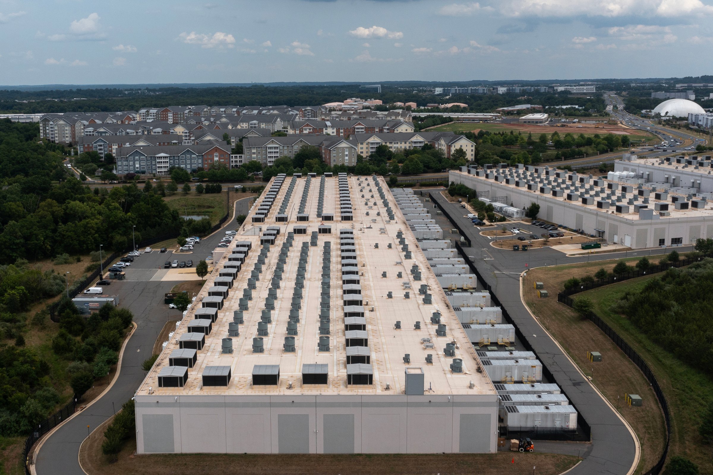 Aerial view of a large commercial building with rows of rooftop mechanical units beside a multifamily residential community.