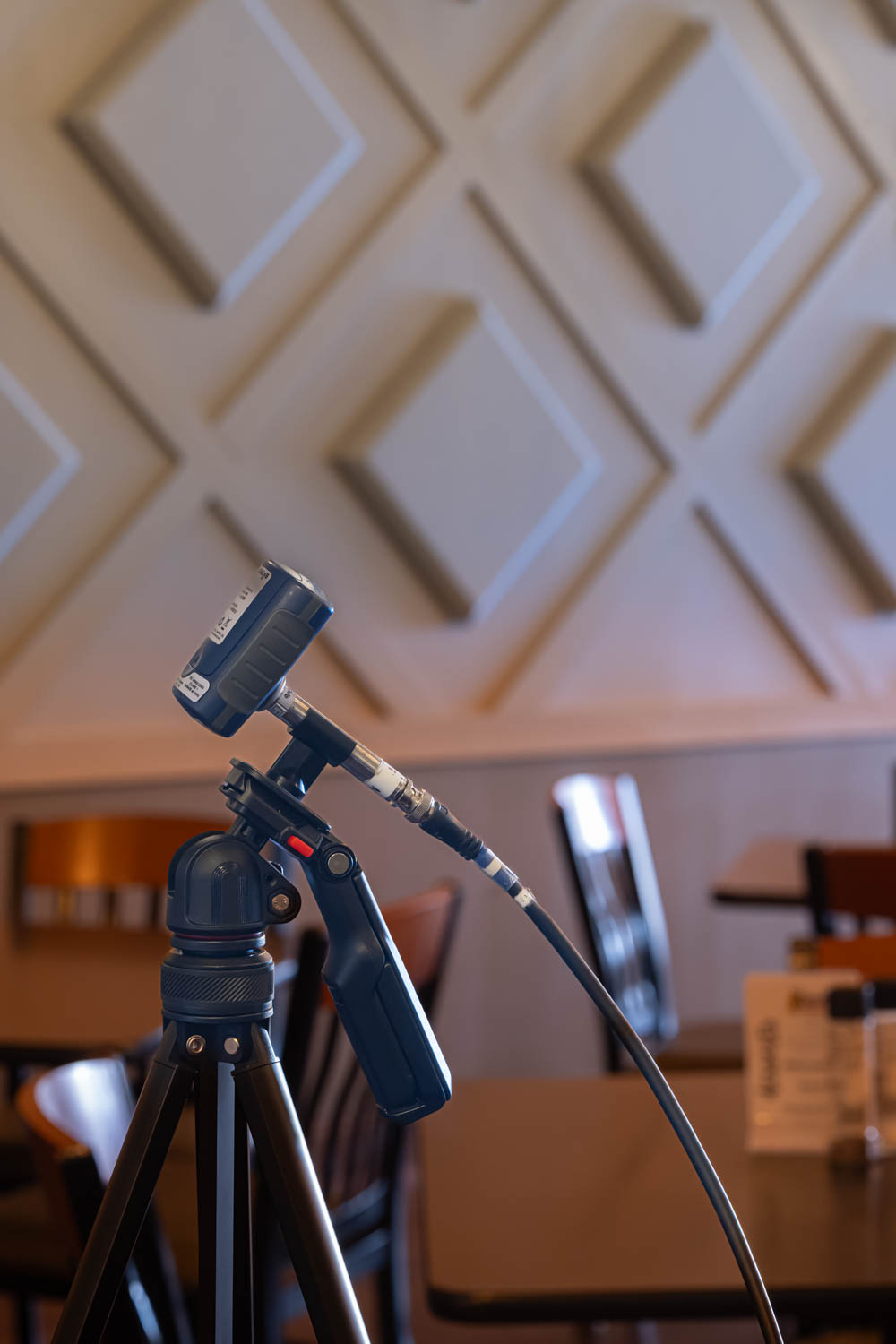 Close-up of a camera device mounted on a tripod with a cable attached, indoors with blurred chairs and tables in the background.