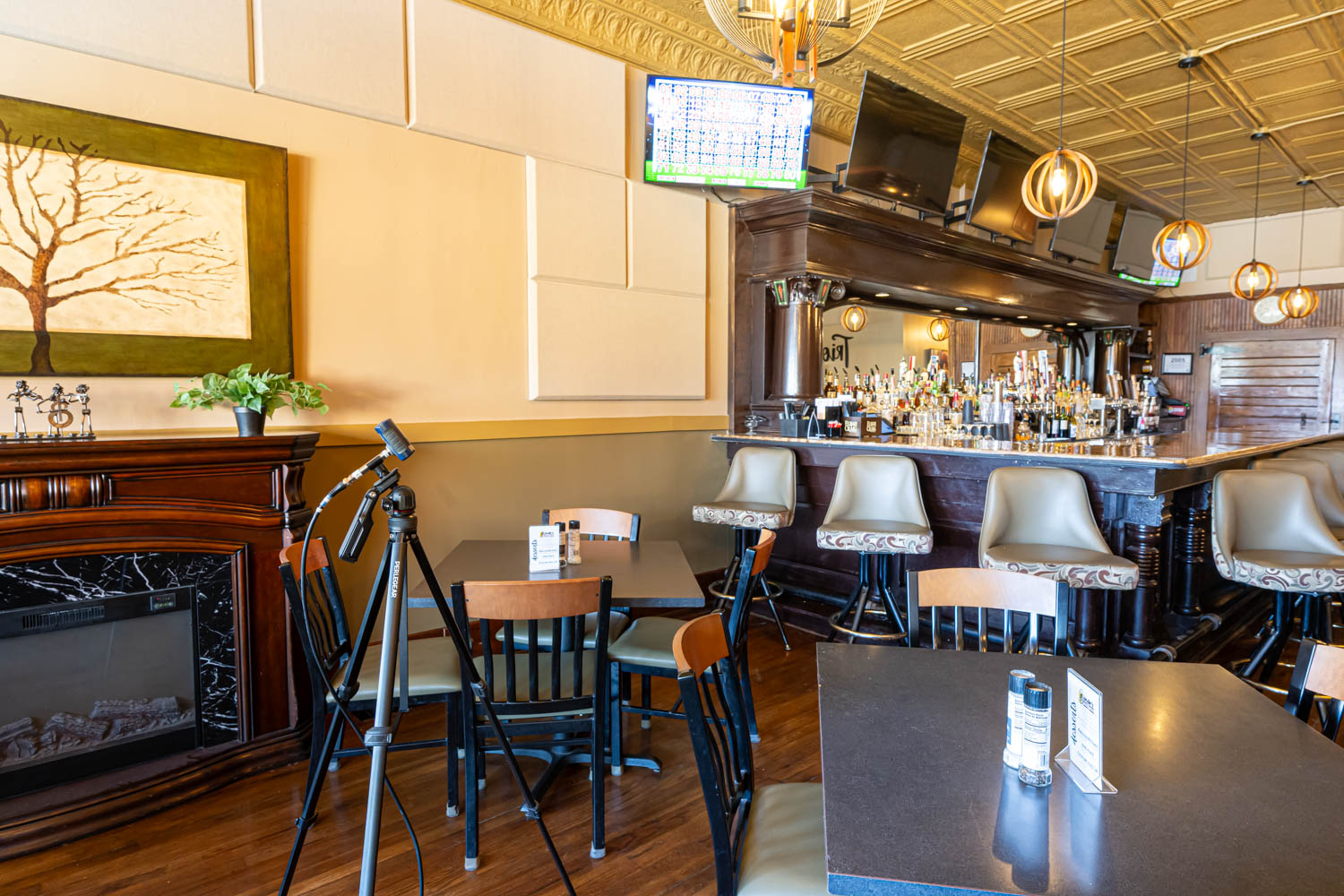 Cozy bar interior with a wooden bar counter lined with cushioned bar stools, tables and chairs in the foreground, a fireplace with a decorative tree painting above it, and a tripod-mounted microphone stand near a table. Pendant lights hang from the ceiling and multiple TVs are mounted above the bar.
