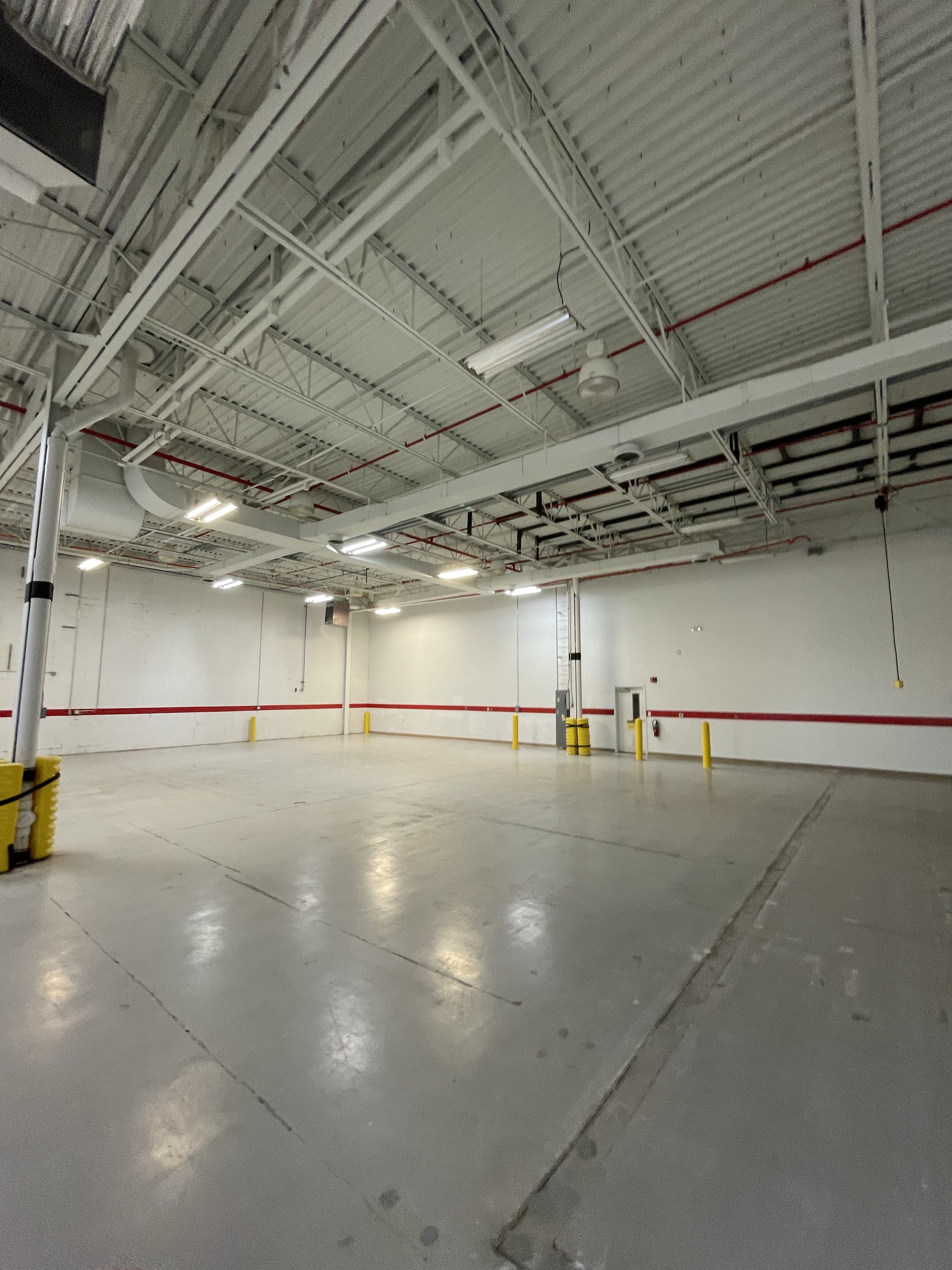 Empty industrial warehouse with high ceilings, white walls marked by a red horizontal stripe, exposed metal beams, overhead fluorescent lighting, and concrete floor with yellow protective guards on support columns.