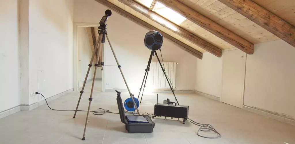 Empty attic room with slanted wooden ceiling beams and beige tiled floor, containing audio measurement equipment including two black tripods with spherical devices, an open black equipment case, and cables on the floor.