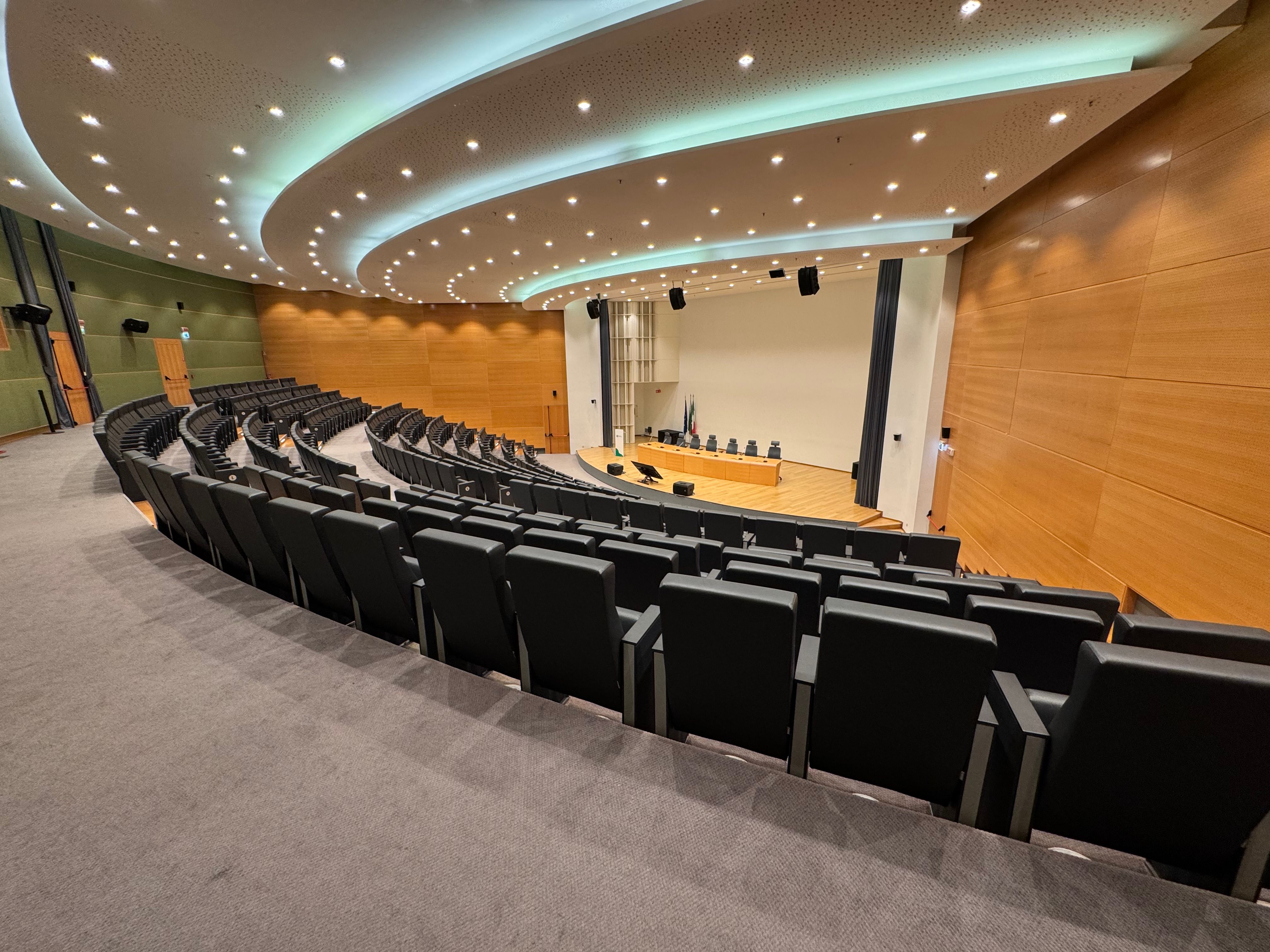 Empty modern lecture hall with curved rows of black chairs facing a stage with a long desk, microphones, monitors, and wooden panel walls beneath a ceiling with circular light patterns.