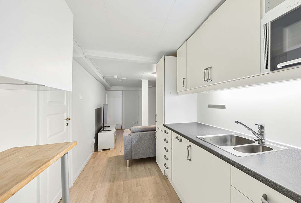 Kitchen remodel in Houston with white cabinets, gray countertop, double sink, and a small living area with gray sofa and TV in the background.