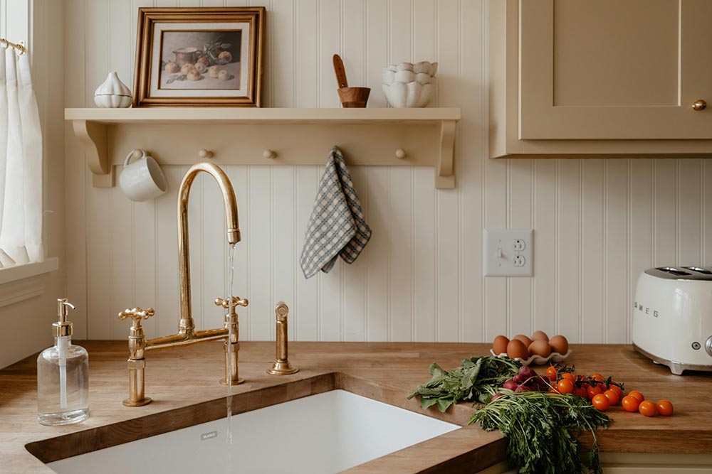 Rustic kitchen sink area with wooden countertop, brass faucet, fresh vegetables, eggs, and a white toaster.