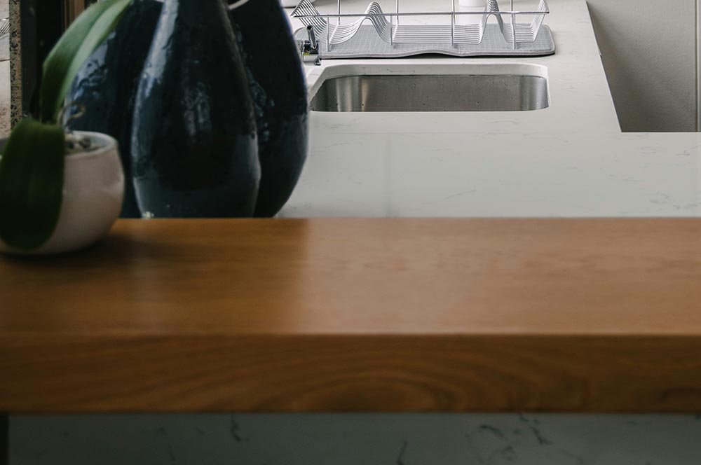 Modern kitchen countertop with a marble sink and wooden bar in the foreground, featuring decorative vases and a small potted plant.