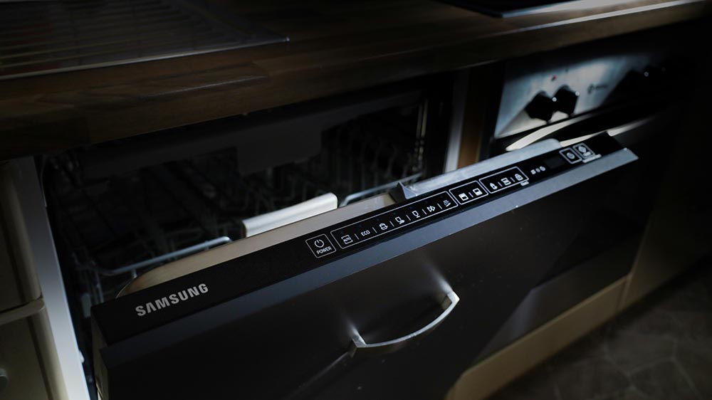 Partially open Samsung dishwasher with illuminated control panel in a kitchen.