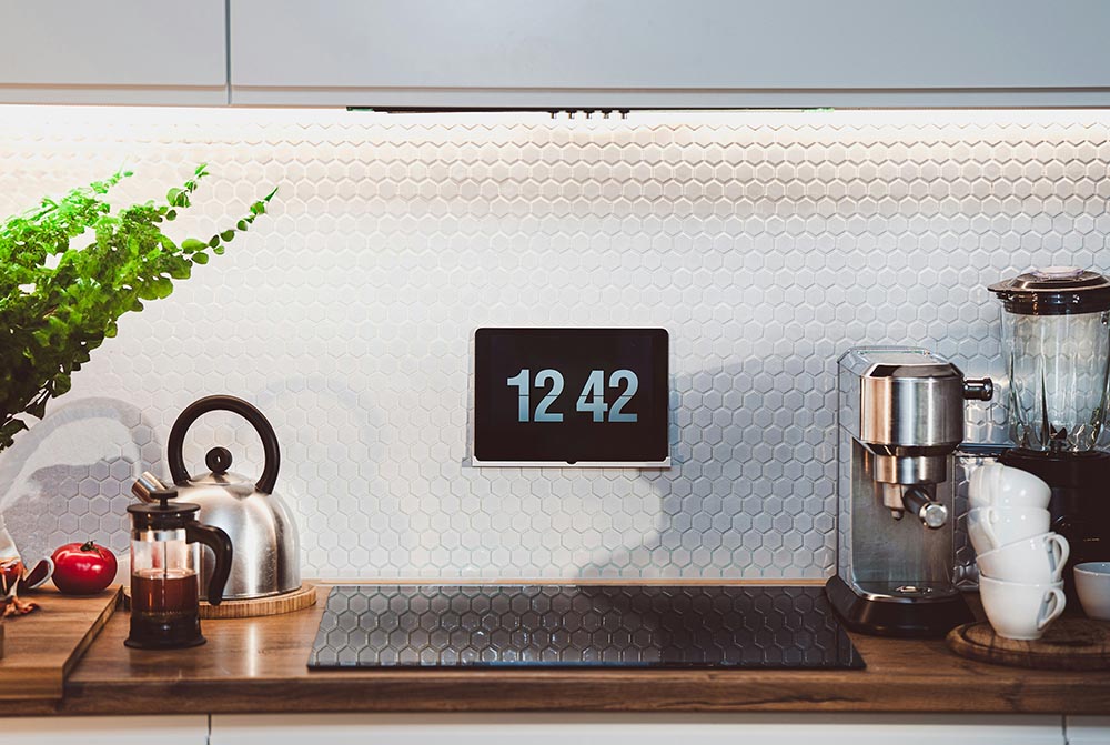 Modern kitchen counter with a wall-mounted digital clock showing 12:42, a kettle, coffee press, coffee machine, blender, stacked white mugs, and a leafy green plant.