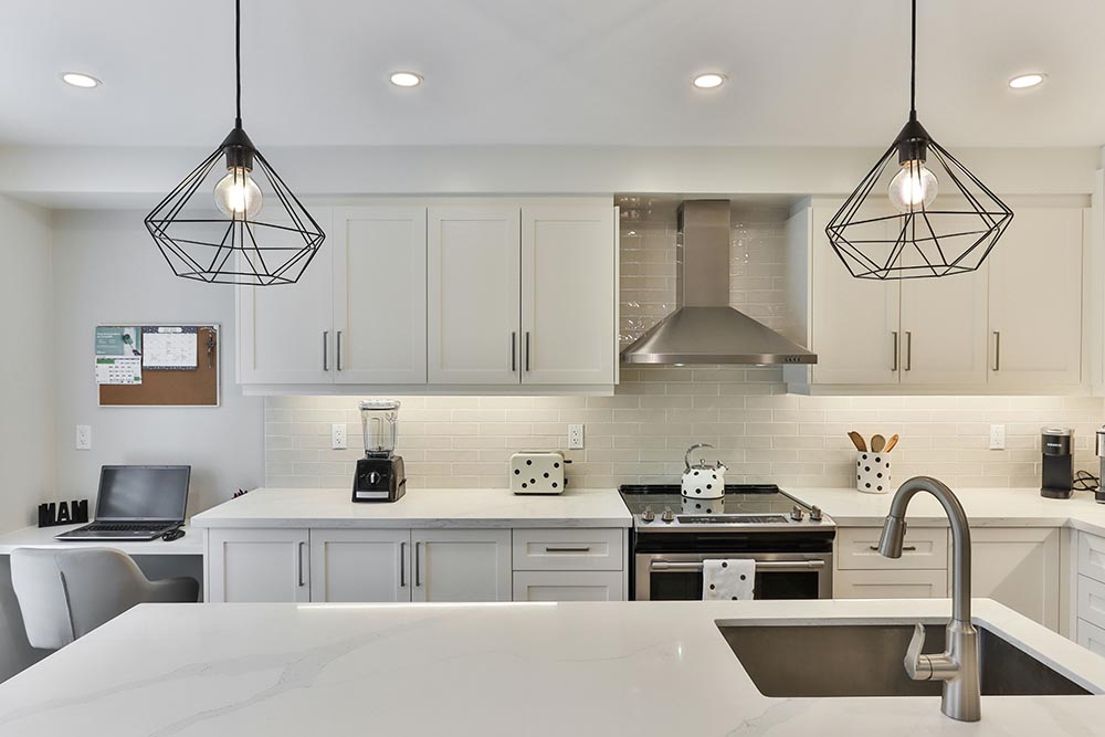 Kitchen remodel in New York with stainless steel stove, range hood, black wireframe pendant lights, white cabinets, and a white countertop with built-in sink.