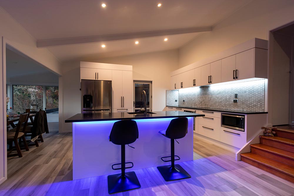 Kitchen remodel in Houston with white cabinets, black countertops, stainless steel refrigerator, under-cabinet lighting, and blue LED lighting on island with two black stools.
