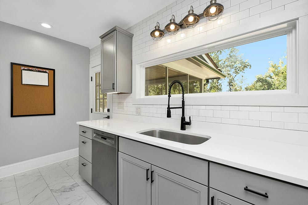 Kitchen remodel in Houston with gray cabinets, white countertop, black single-handle spring coil faucet, large window showing trees outside, and subway tile backsplash.