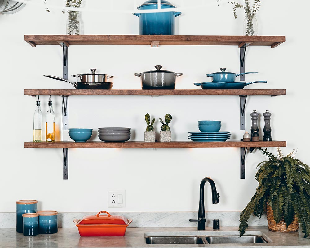 Three wooden kitchen shelves mounted on a white wall holding pots, pans, bowls, plates, and decorative plants above a marble countertop with a double sink and black faucet, next to a green fern in a wicker basket.