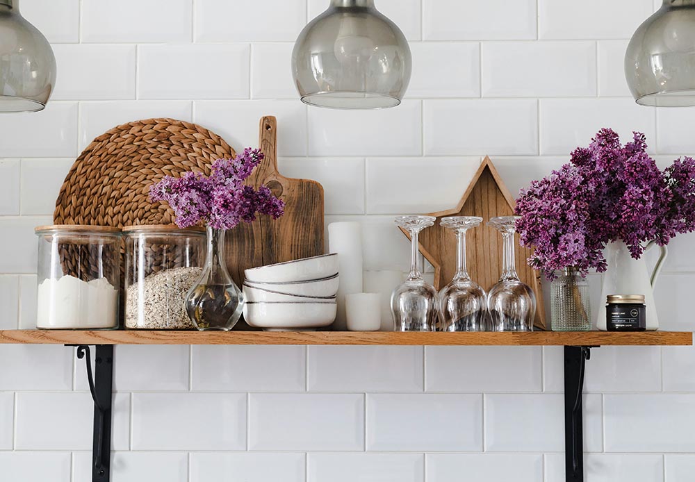 Wooden kitchen shelf with glass jars of flour and oats, purple flowers in glass vases, white bowls, upside-down wine glasses, a star-shaped wooden decor, and a small candle against white tiled wall.