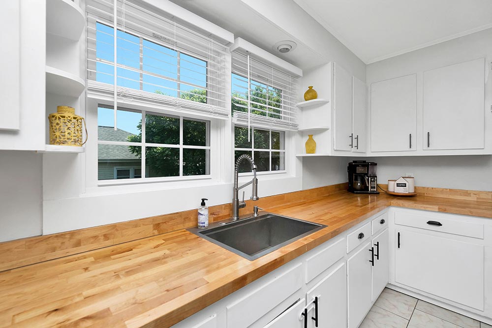 Bright Kitchen remodel in Chicago with wooden countertops, white cabinets, a black sink under two large windows with white blinds, and a coffee maker on the corner counter.
