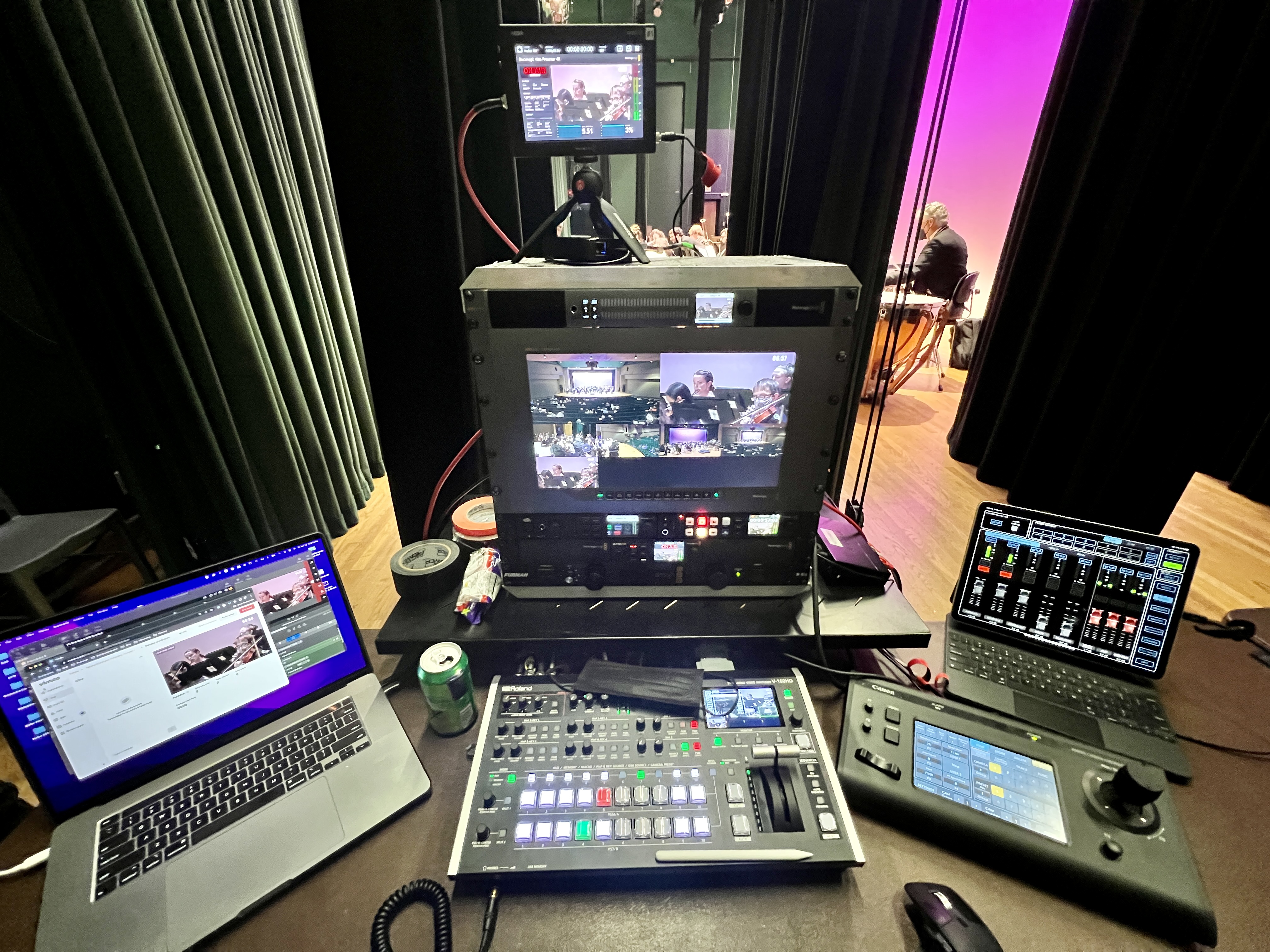 Backstage livestream production rack with switcher, multiview monitor, and audio mixer with stage visible in background