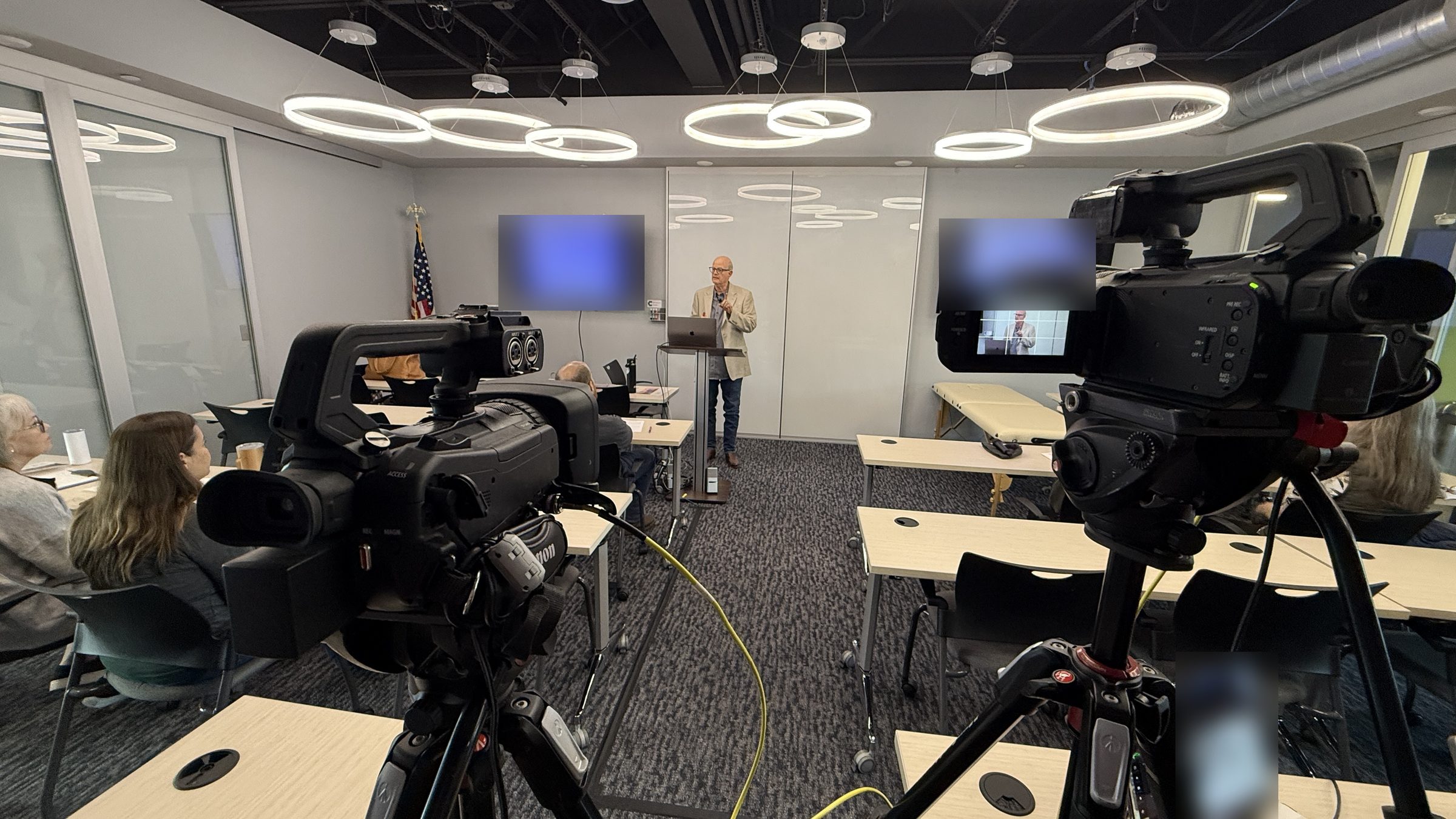 Two cameras covering a presenter in a corporate classroom with a small in-person audience while the session streams to remote attendees