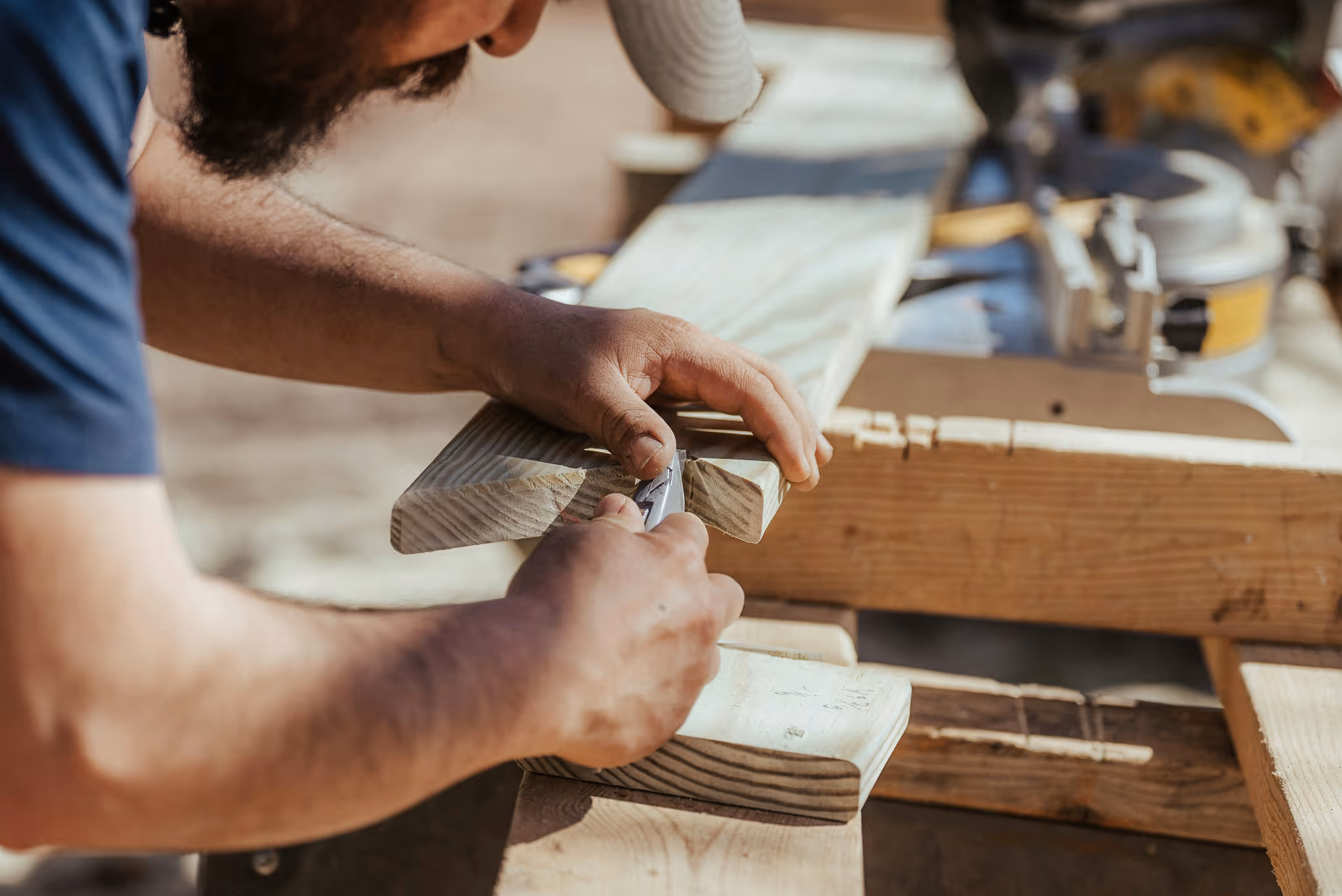 Close-up of a craftsman carving or shaving a piece of timber with a utility knife, working on a wooden joint at an outdoor woodworking station.