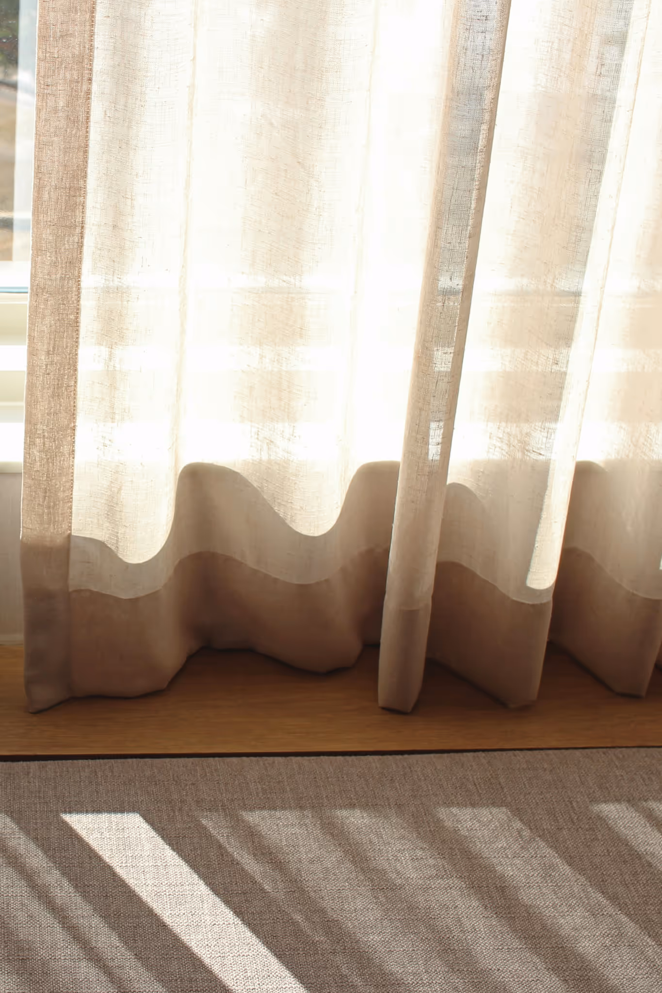 Close-up of sheer beige linen curtains with sunlight streaming through, casting shadows on a textured carpet floor.