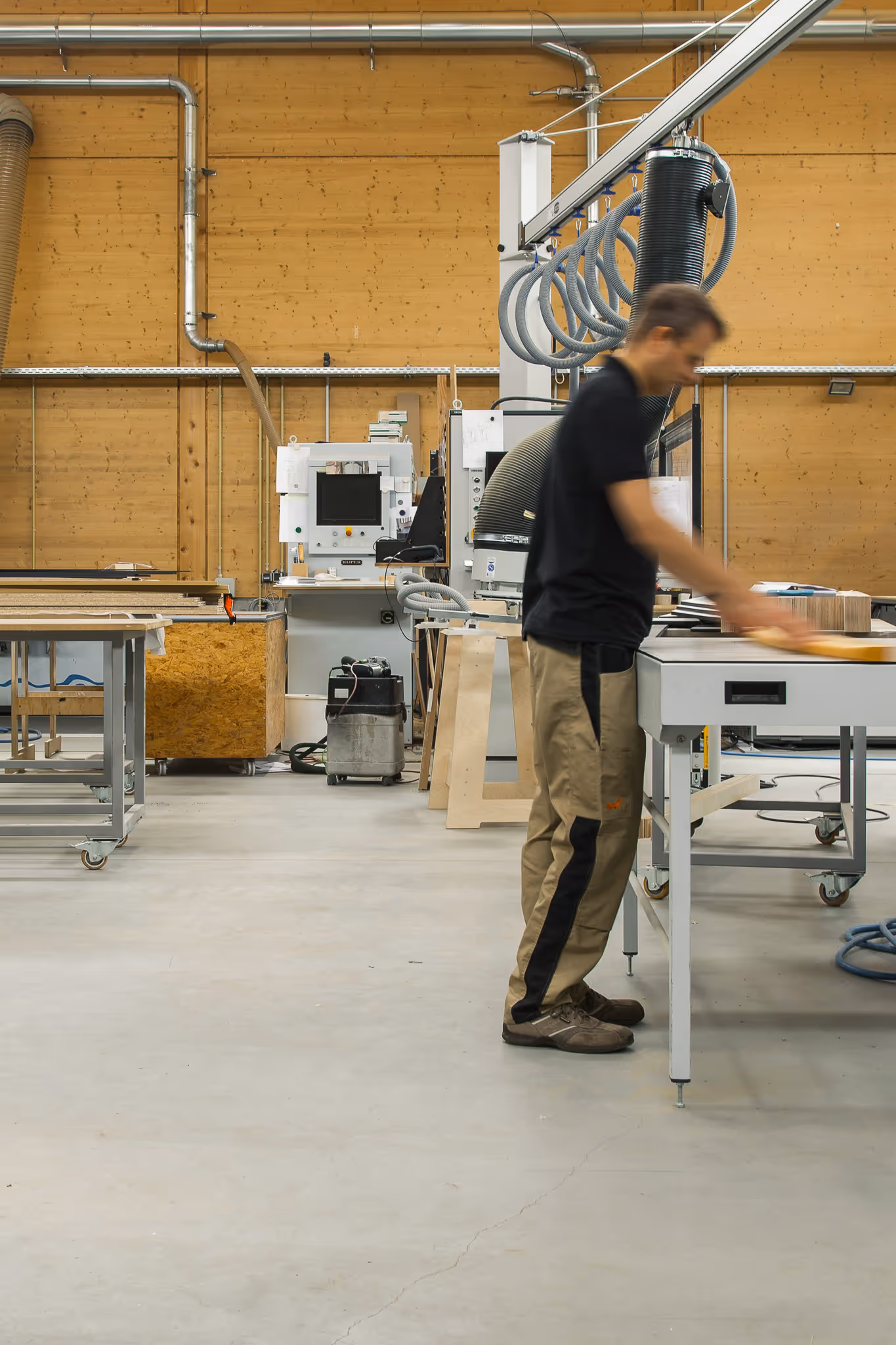 Woodworking workshop with craftsman sanding wood at workbench, surrounded by modern equipment and wood-paneled walls.