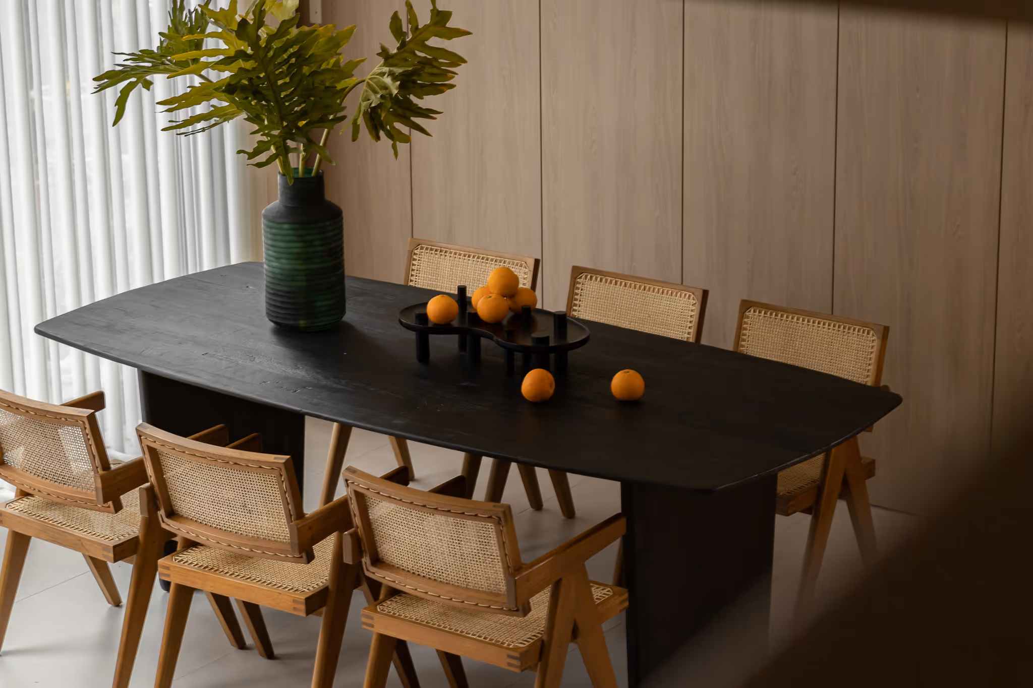Modern dining room with a dark wooden table, natural rattan chairs, a green striped vase with leafy branches, and fresh oranges arranged as decor.