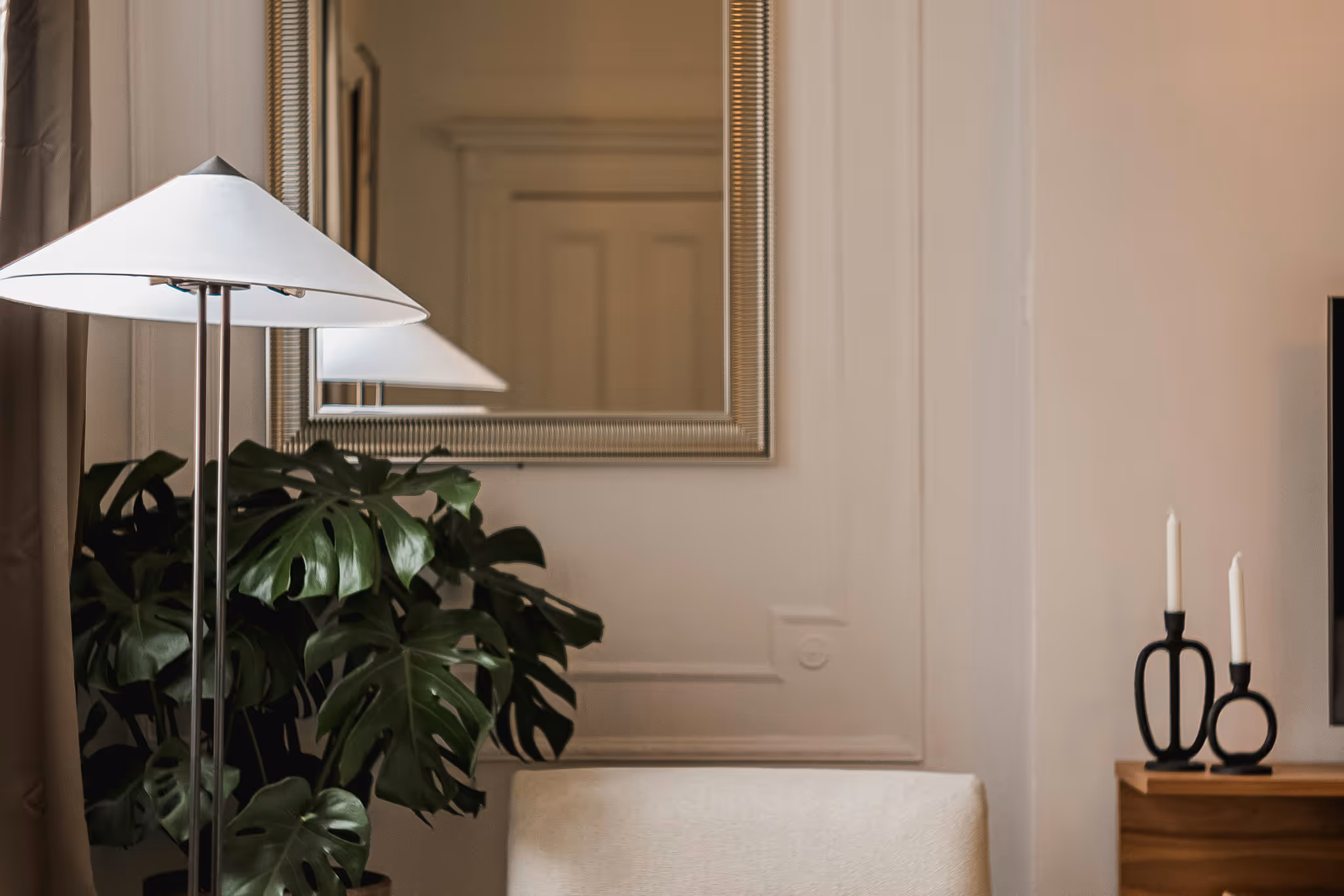 Cozy living room corner with white floor lamp, large mirror, Monstera plant, cream armchair, and decorative candles.