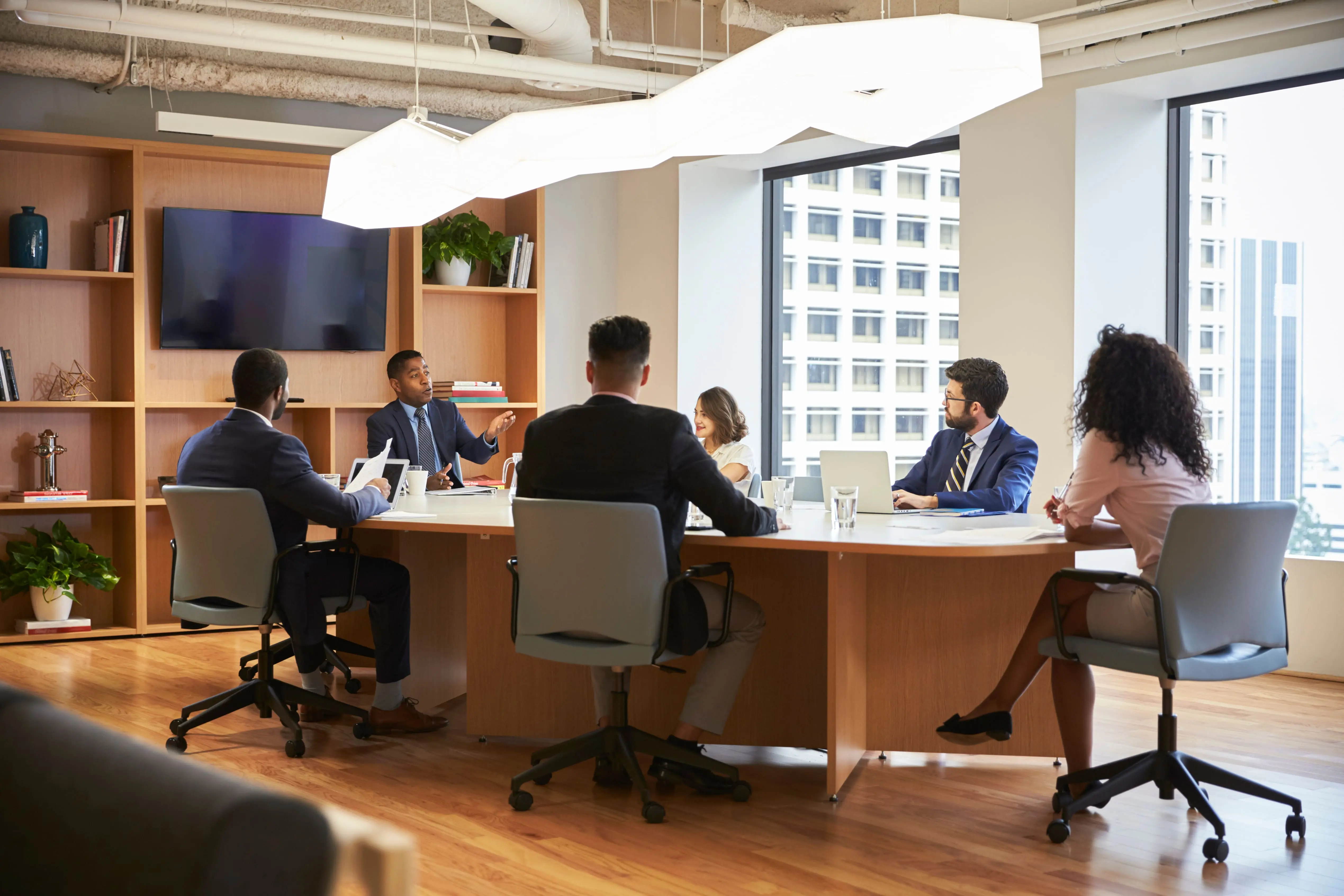 Five professionals in business attire engaged in a meeting around a wooden conference table in a modern office with large windows and bookshelves.