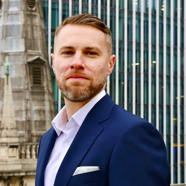 Man with short hair and beard wearing a navy blue suit and white shirt, standing outdoors in front of modern and historic buildings.