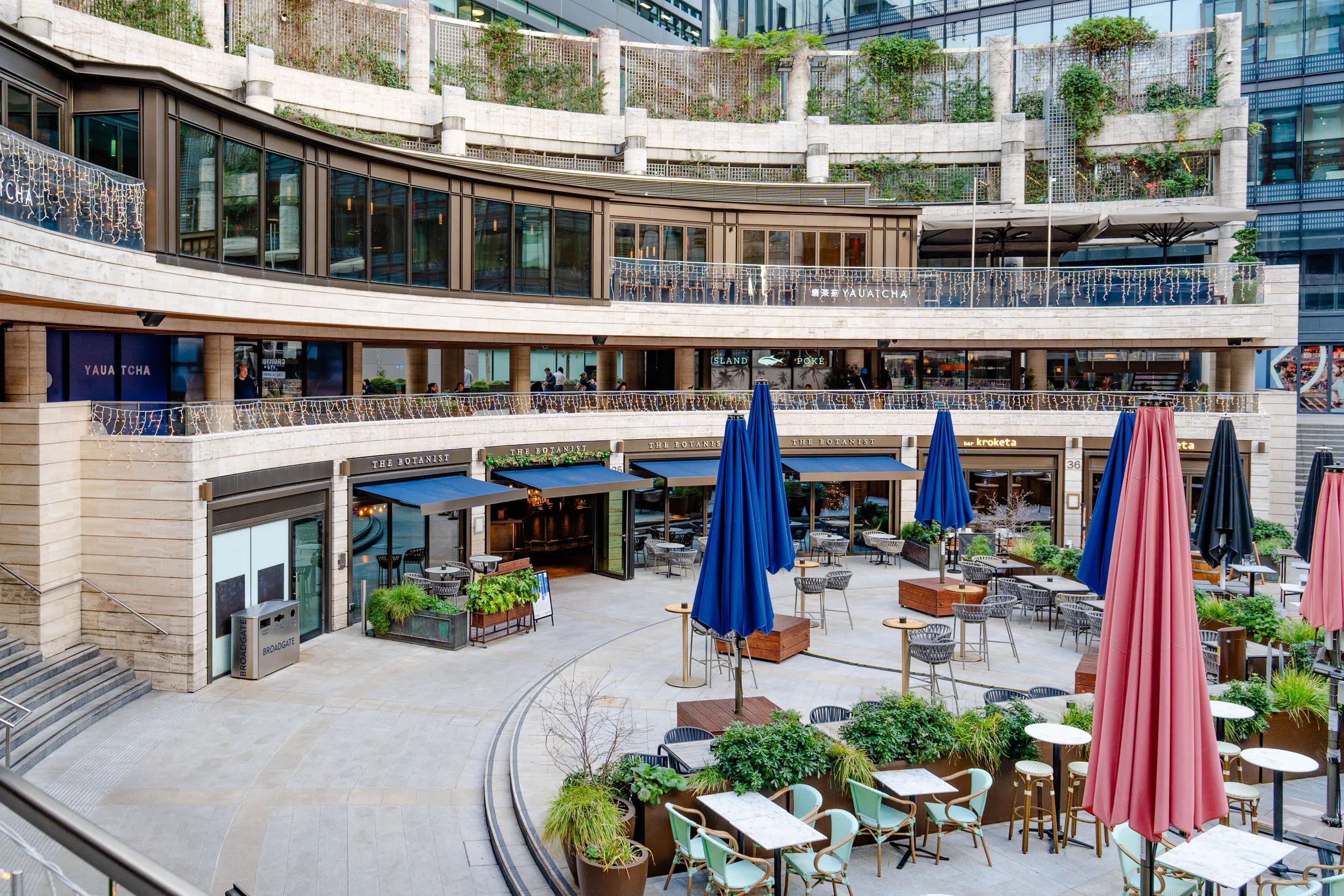 Outdoor dining area with multiple tables, chairs, and closed umbrellas in front of restaurants in a modern curved building.