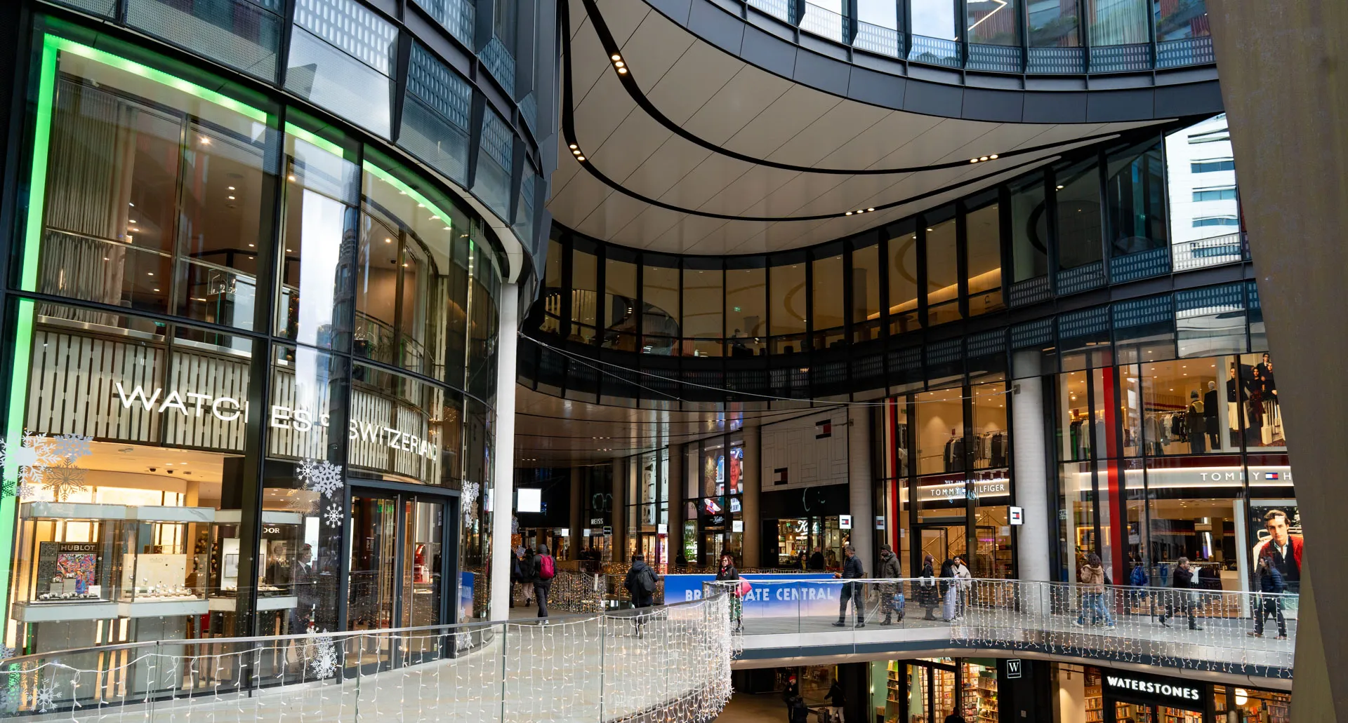 Interior of a modern shopping mall with stores including Watches of Switzerland, Tommy Hilfiger, and Waterstones, featuring glass facades and people walking.