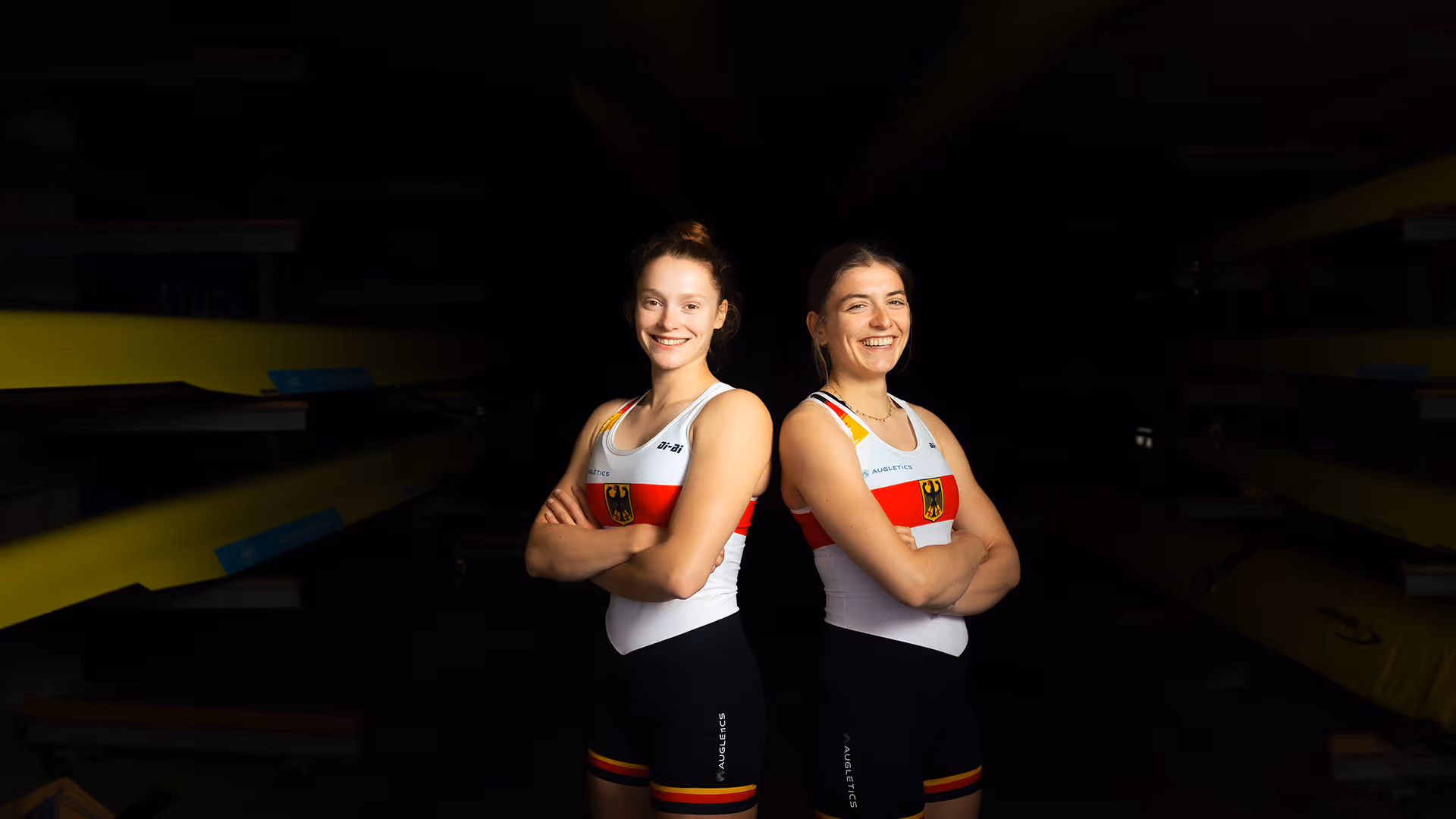 Porträtfoto von zwei Ruderinnen im Einteiler des deutschen Nationalteams, stehend nebeneinander mit verschränkten Armen in einer Bootshalle.