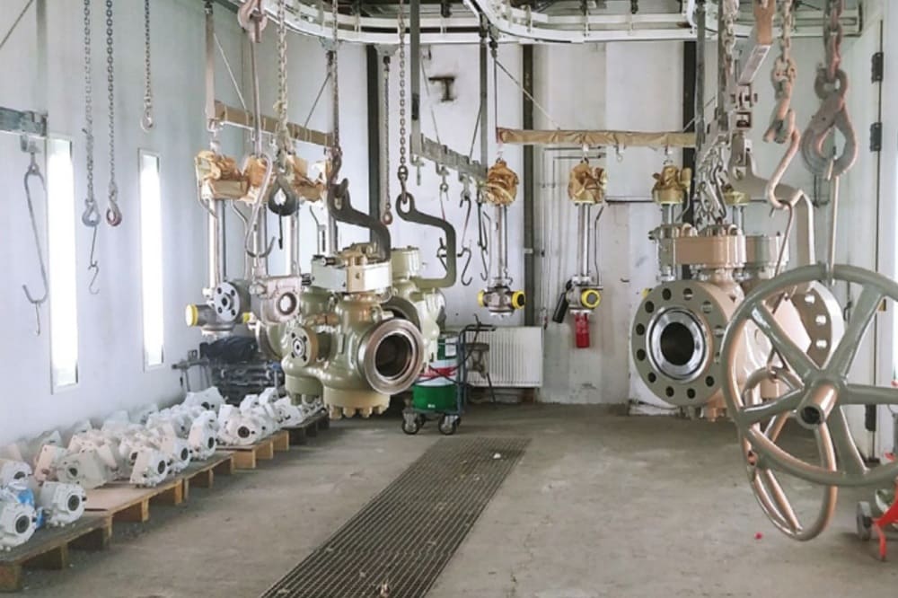 Industrial workshop with various large metal valves and valve components hanging from chains and lined on pallets.